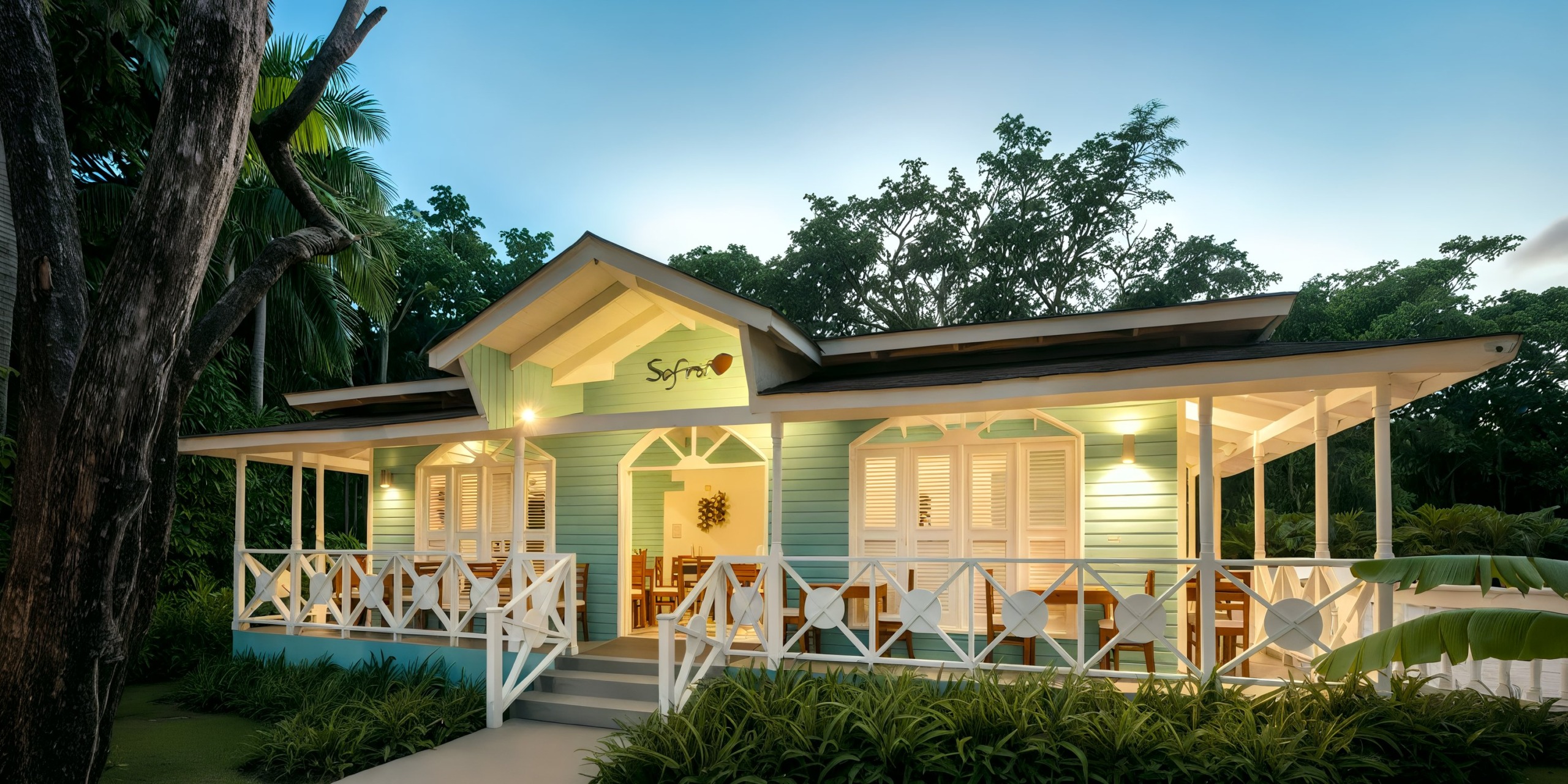 A cozy, pastel-colored bungalow nestled in lush greenery at the Viva Wyndham V Heavens - All-Inclusive Resort, Adults Only. At dusk, the wraparound porch is adorned with wooden furniture and warmly illuminated lights.