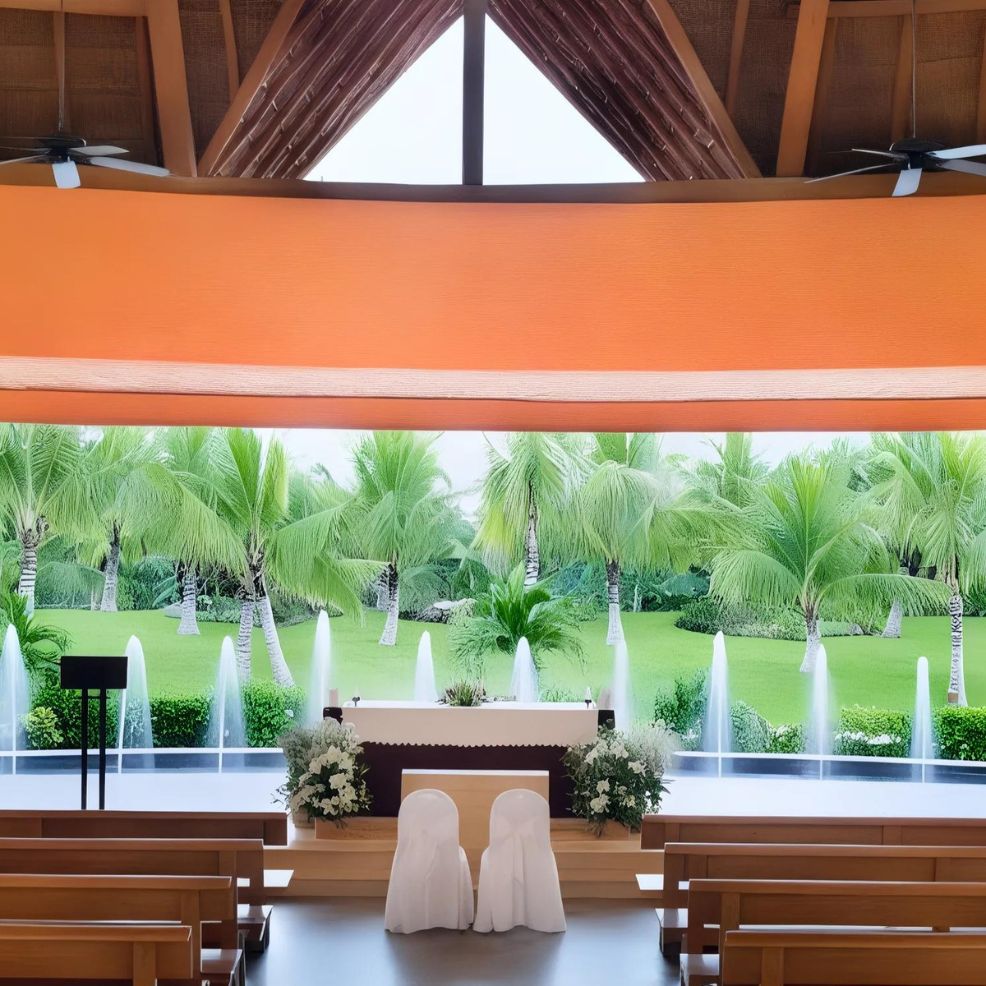 The interior of Barcelo Maya Riviera features wooden benches, a beautifully decorated altar, and expansive windows offering views of palm trees and fountains.