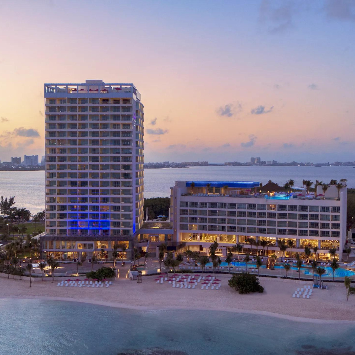 Aerial view of Breathless Cancún Soul Resort & Spa at dusk, ideal for destination weddings, featuring illuminated buildings and a pool area, with a city skyline in the background.