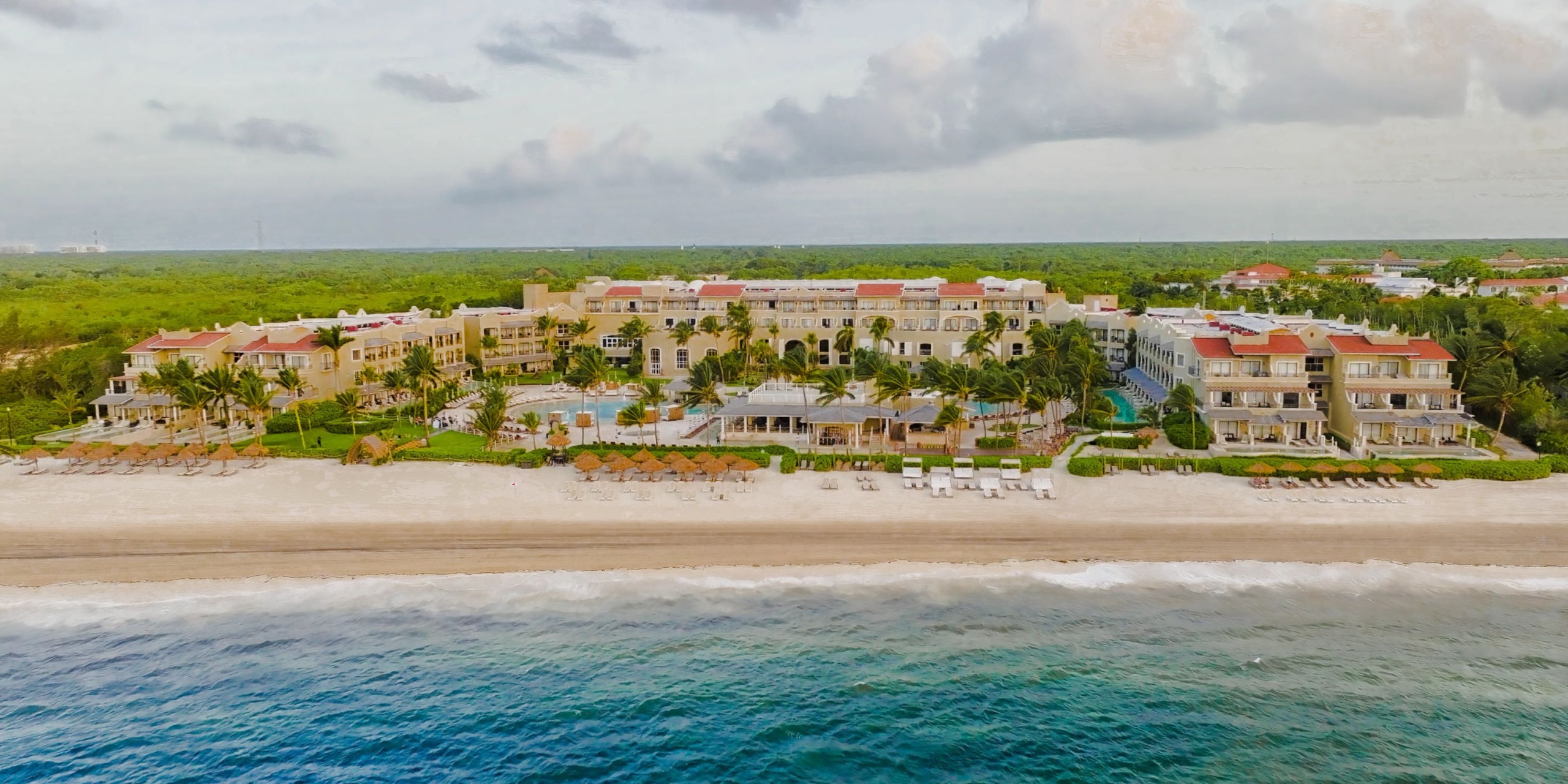 An aerial view of Hyatt Zilara Riviera Maya, a beachfront resort featuring multiple buildings, a large pool, sun loungers, and palm trees, all set against a backdrop of dense greenery and an overcast sky.