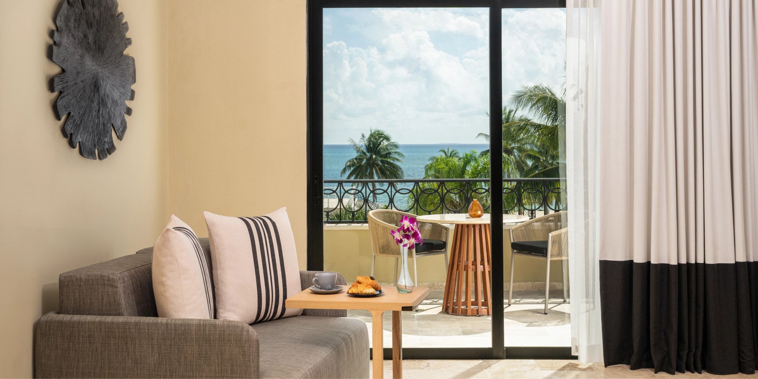 A living room at the Hyatt Zilara Riviera Maya features a sofa, coffee on a table, and an ocean-view balcony. Outside, there's a table with chairs and a potted plant. White and black curtains frame the sliding glass doors.