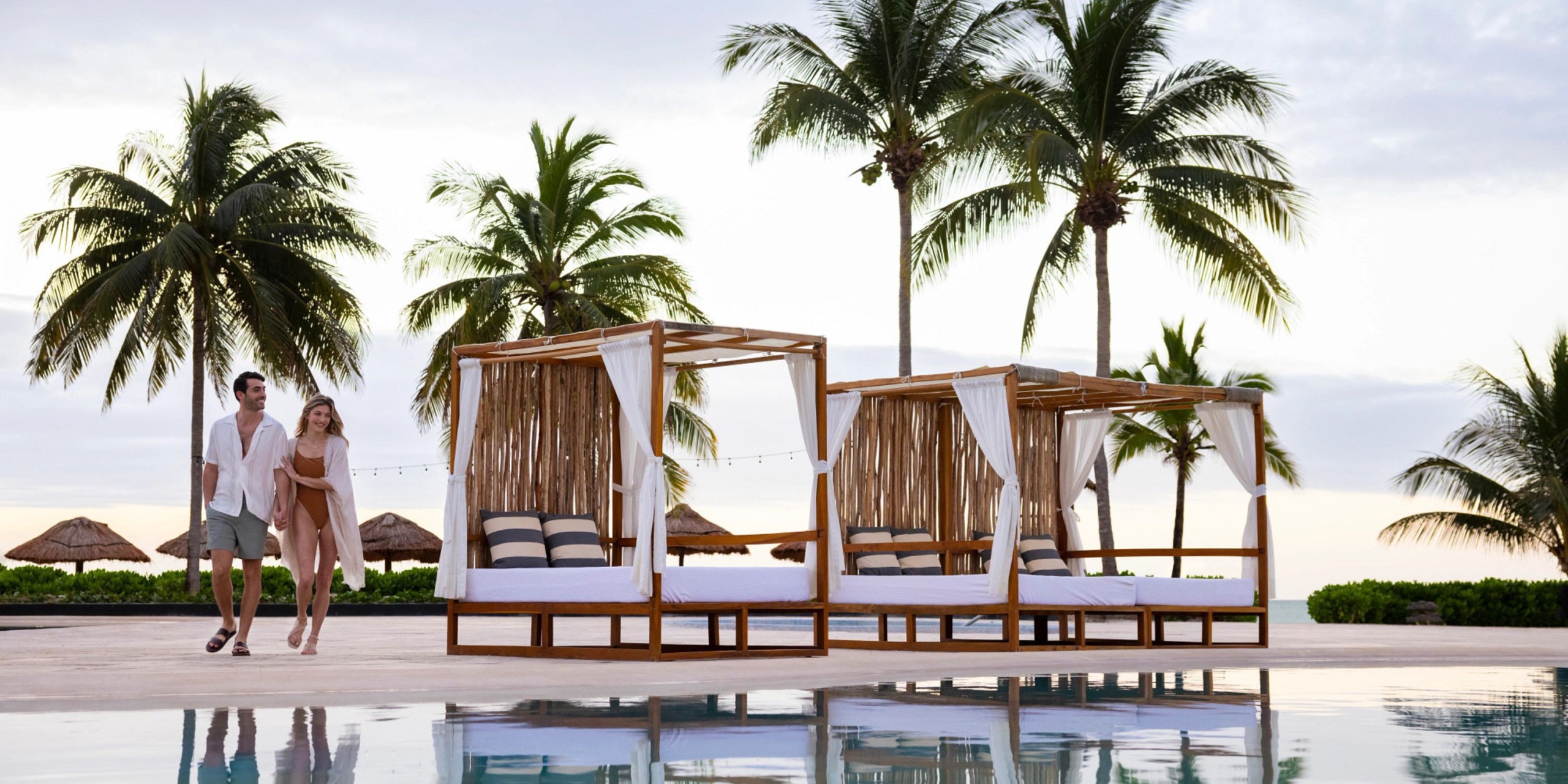 A couple strolls by a pool bordered with cabanas and palm trees at the Hyatt Zilara Riviera Maya.