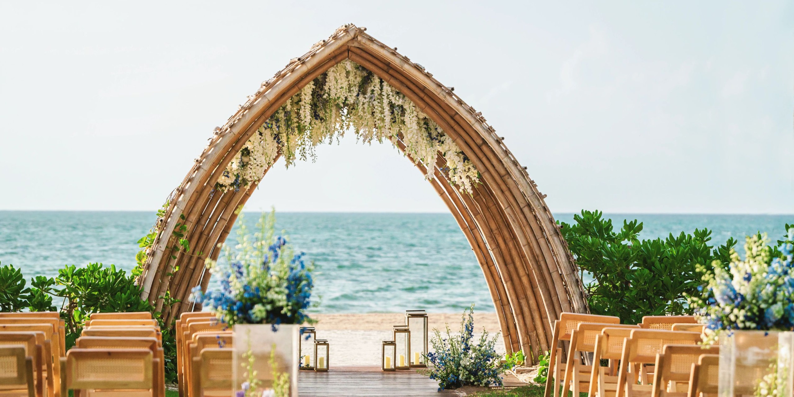 Experience a beach wedding setup at the Hyatt Zilara Riviera Maya, featuring a stunning floral arch crafted from intertwined branches, wooden chairs elegantly arranged, and beautiful blue floral arrangements, all with a breathtaking view of the ocean.