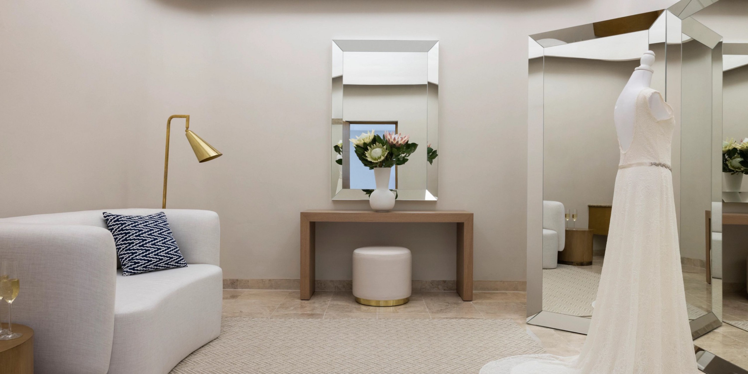 A contemporary fitting room featuring a white dress on a mannequin, complemented by a large mirror, an elegant gold floor lamp, a sleek white sofa adorned with a blue pillow, and accompanied by a chic wooden table with a vase of flowers and an inviting round stool reminiscent of the luxurious ambiance at the Hyatt Zilara Riviera Maya.
