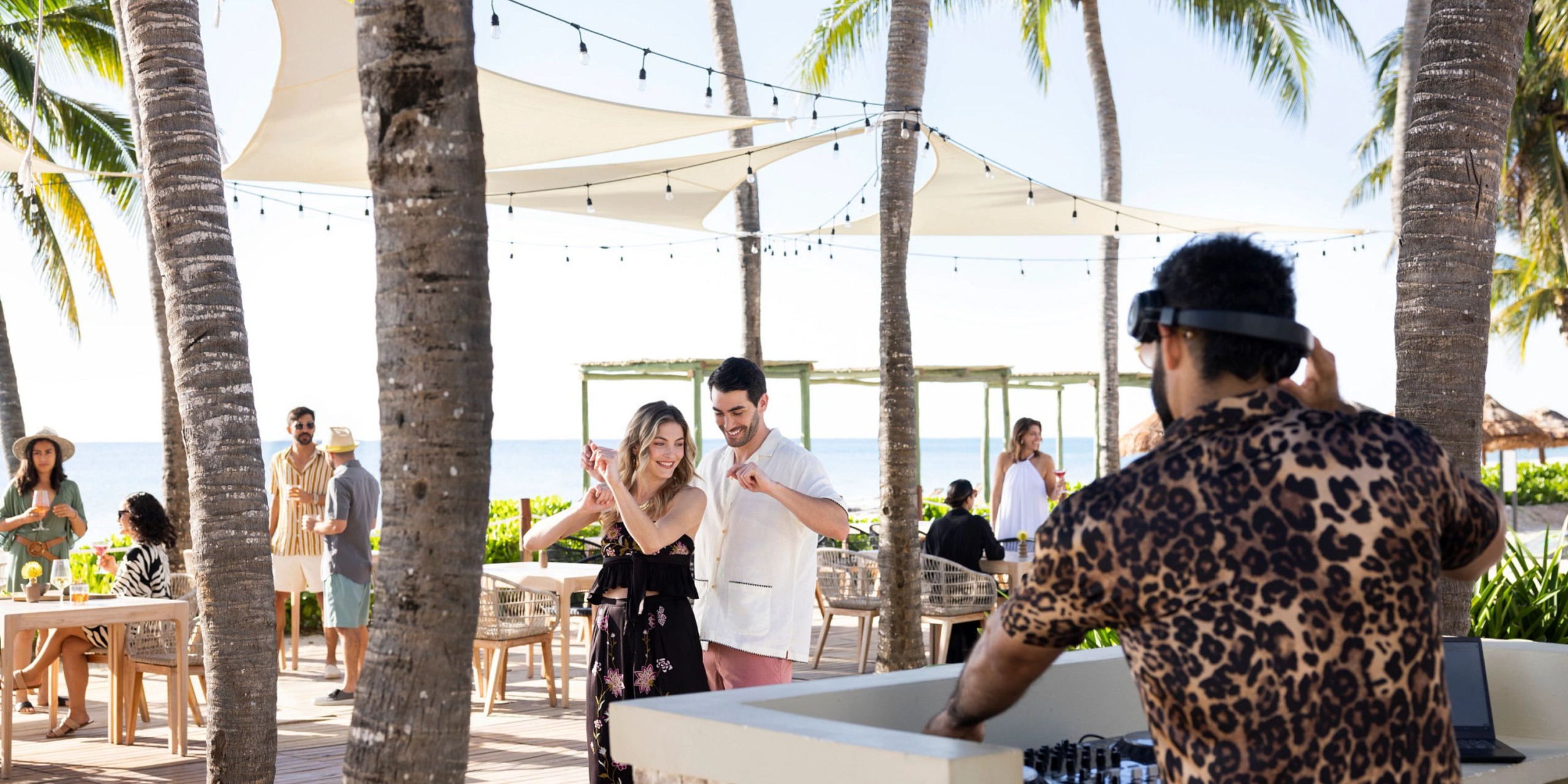 A couple dances near a DJ at the outdoor beachside venue of Hyatt Zilara Riviera Maya, while others sit and stand around, enjoying the atmosphere under palm trees and shaded canopies.