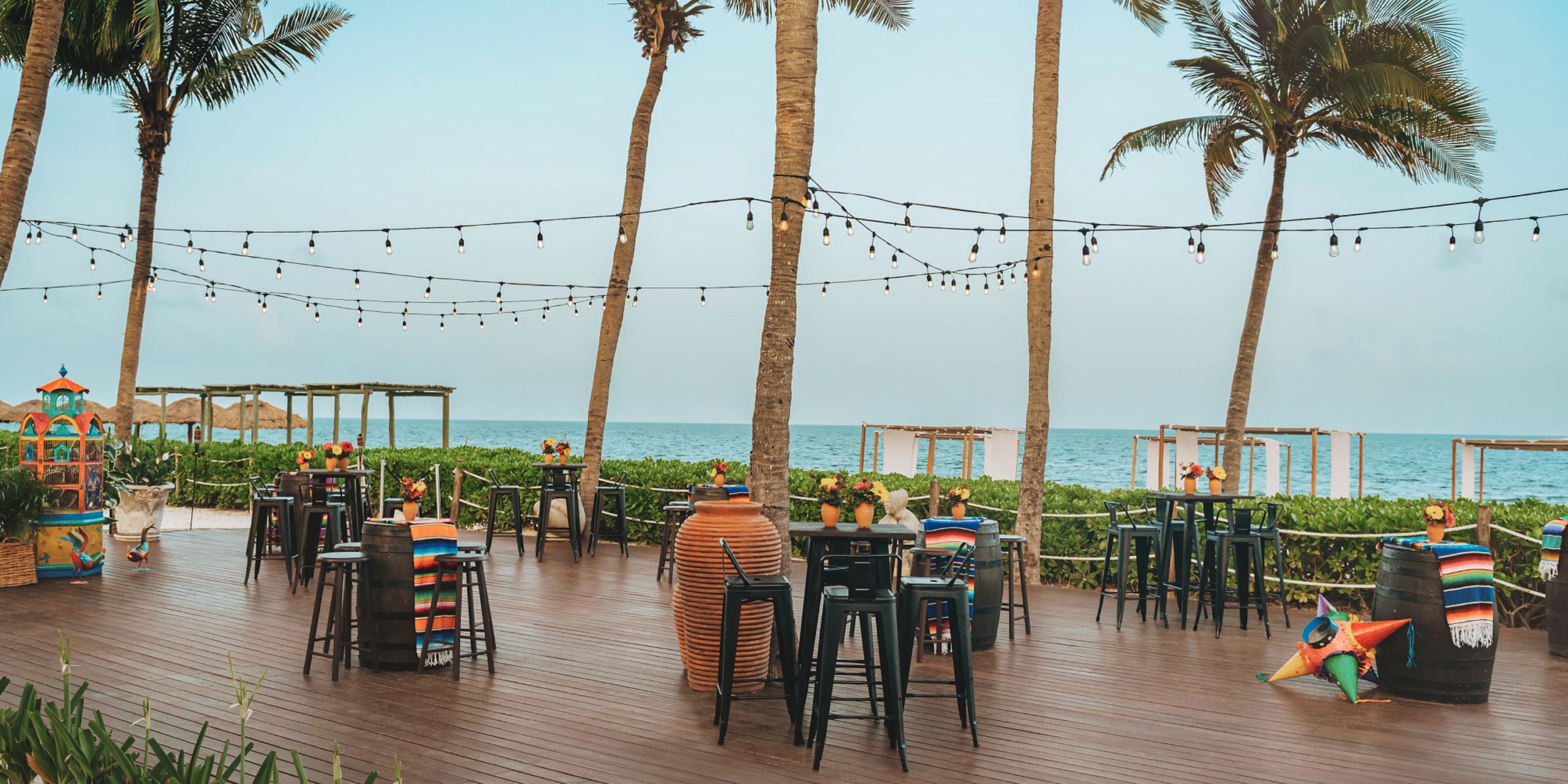 Experience the Hyatt Zilara Riviera Maya's outdoor beachfront deck, featuring tables and stools beneath twinkling string lights, surrounded by swaying palm trees with a backdrop of the ocean and a clear sky.