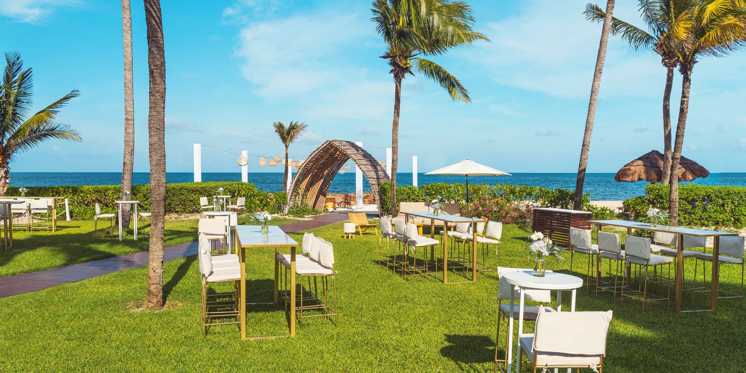 The Hyatt Zilara Riviera Maya offers a stunning outdoor beach wedding setup featuring white chairs and tables, surrounded by palm trees, with a beautiful archway oriented towards the ocean beneath a clear blue sky.