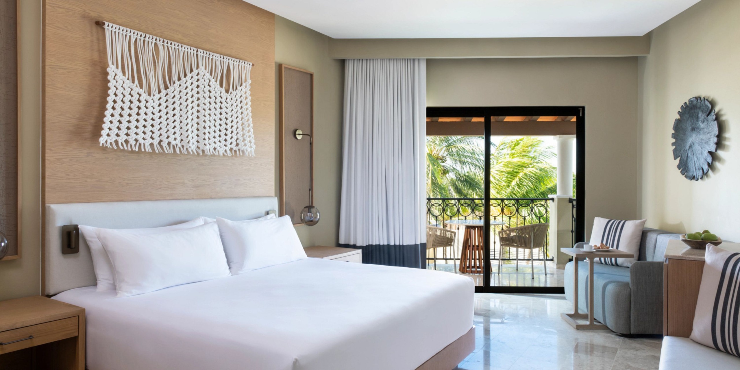 Experience the Hyatt Zilara Riviera Maya, featuring a contemporary hotel room with a king-size bed, elegant macrame wall art, a cozy seating area, and a balcony with breathtaking views of palm trees.