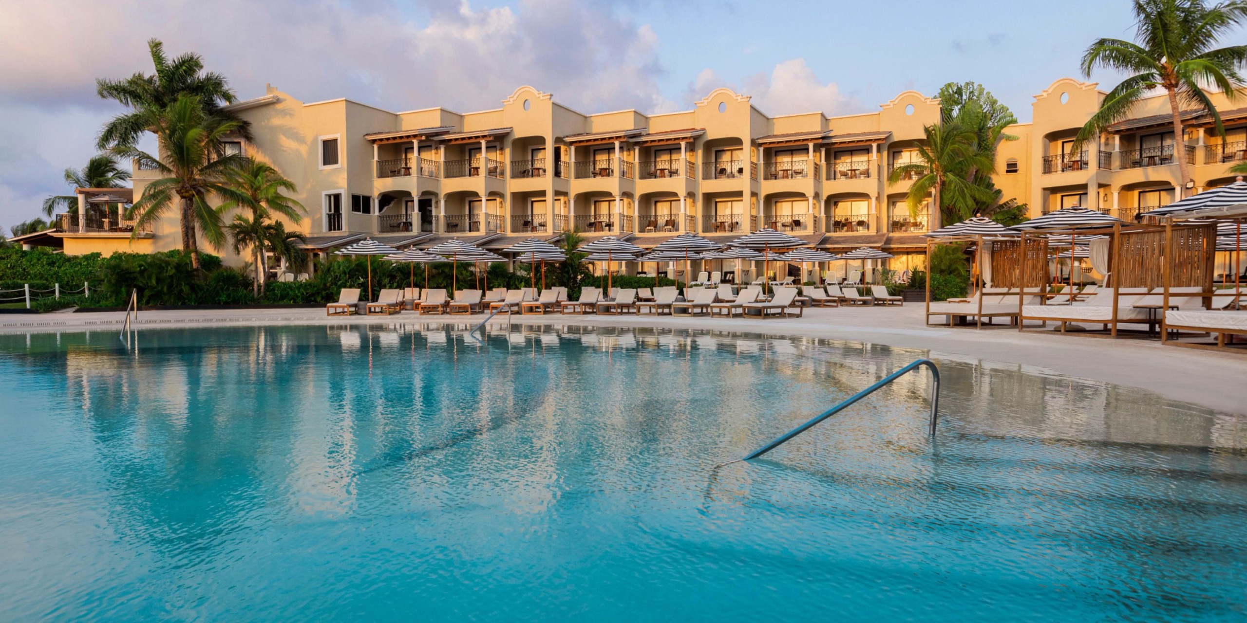 The Hyatt Zilara Riviera Maya features a spacious outdoor swimming pool, complemented by lounge chairs and umbrellas, set against the backdrop of a three-story beige hotel structure encircled by palm trees.