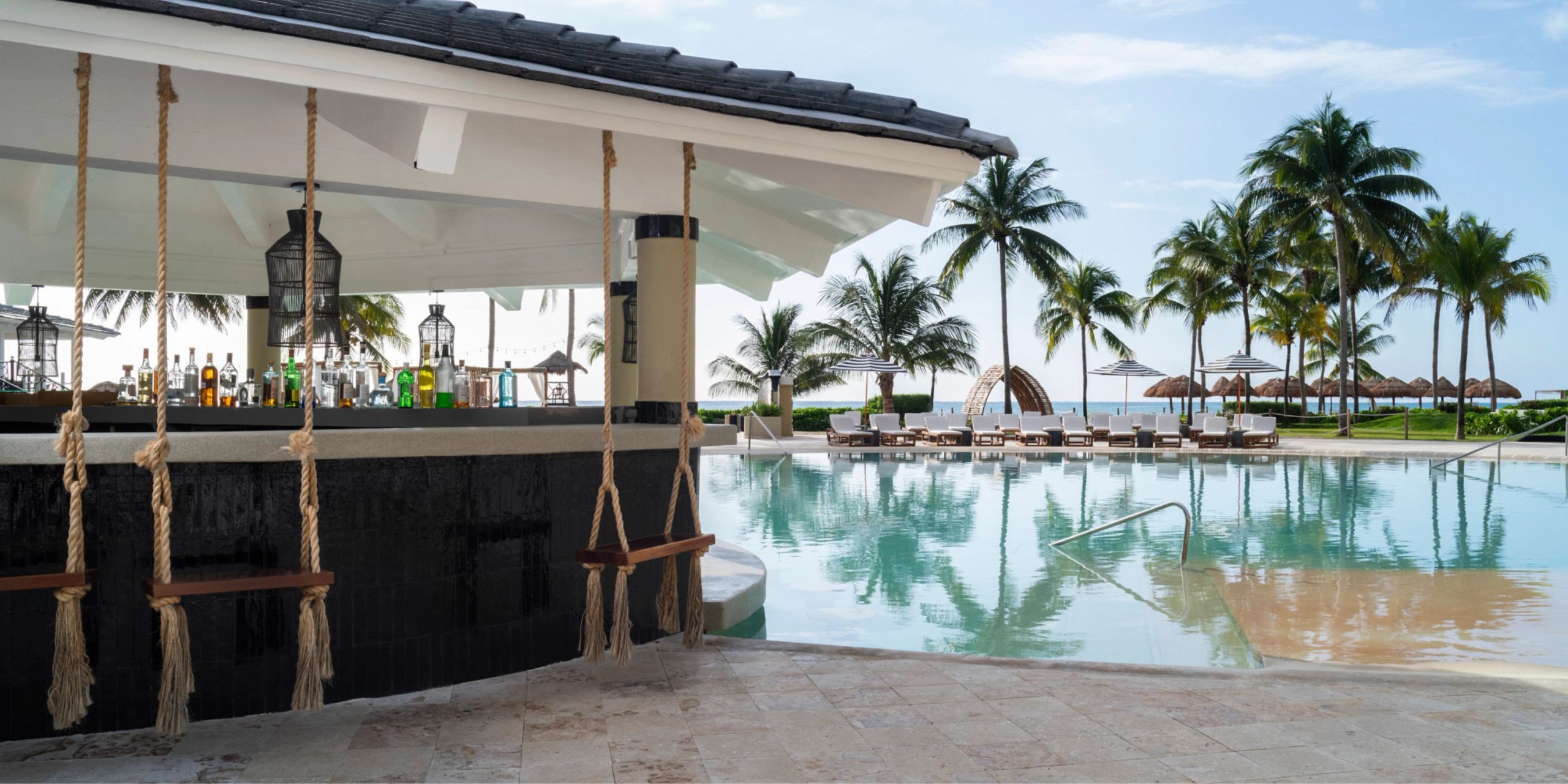 At Hyatt Zilara Riviera Maya, enjoy a poolside bar featuring swings, encircled by palm trees and sun loungers, all with a stunning view of the ocean on a clear day.