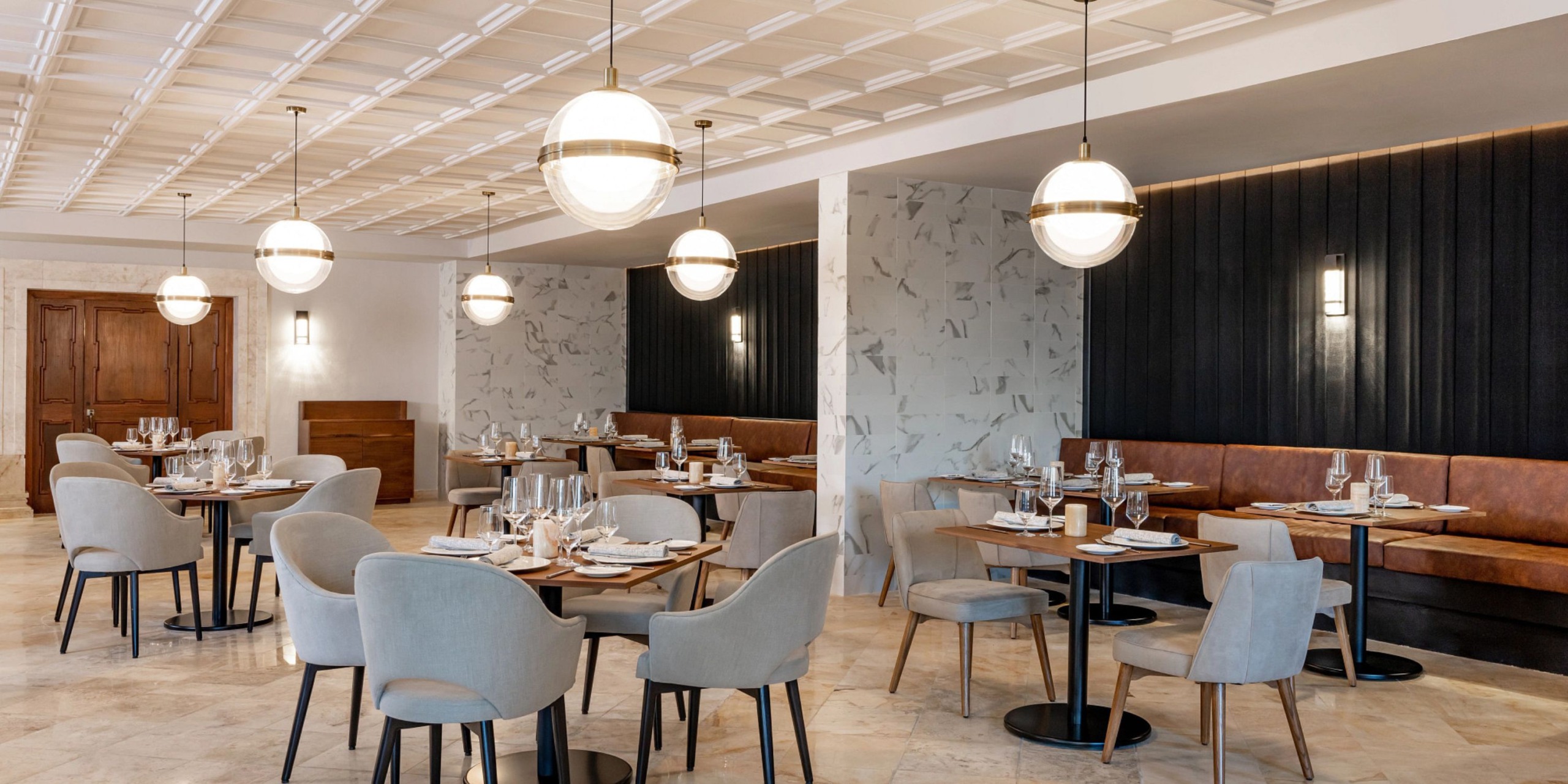 The sophisticated interior of Hyatt Zilara Riviera Maya features round tables, upholstered chairs, and pendant lighting. Tables are elegantly set with glasses and cutlery, complemented by walls that artfully combine white and dark panels.