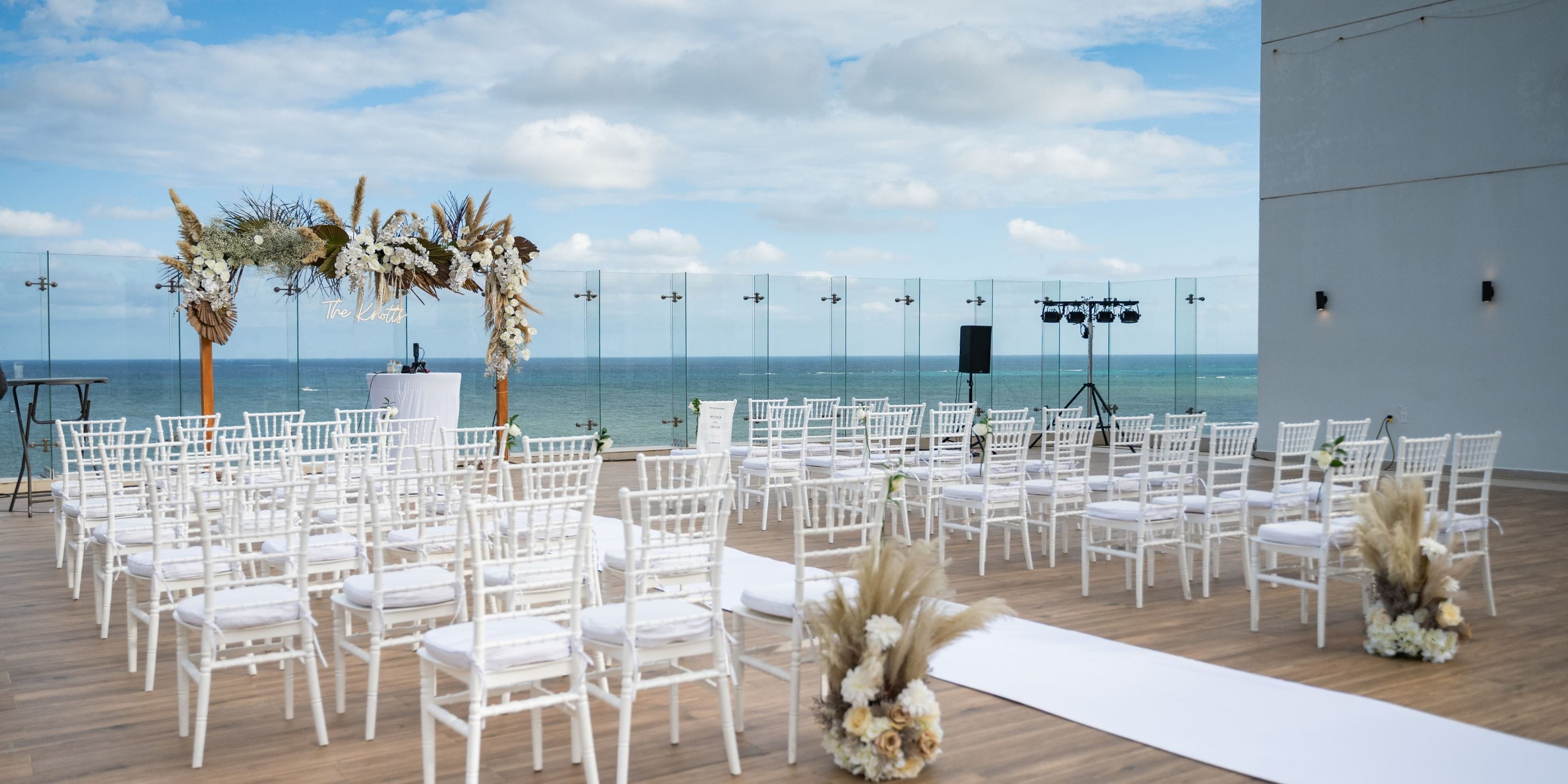 White chairs are arranged for a destination wedding, facing a floral arch on the seaside deck at Royalton Splash Riviera Cancun.