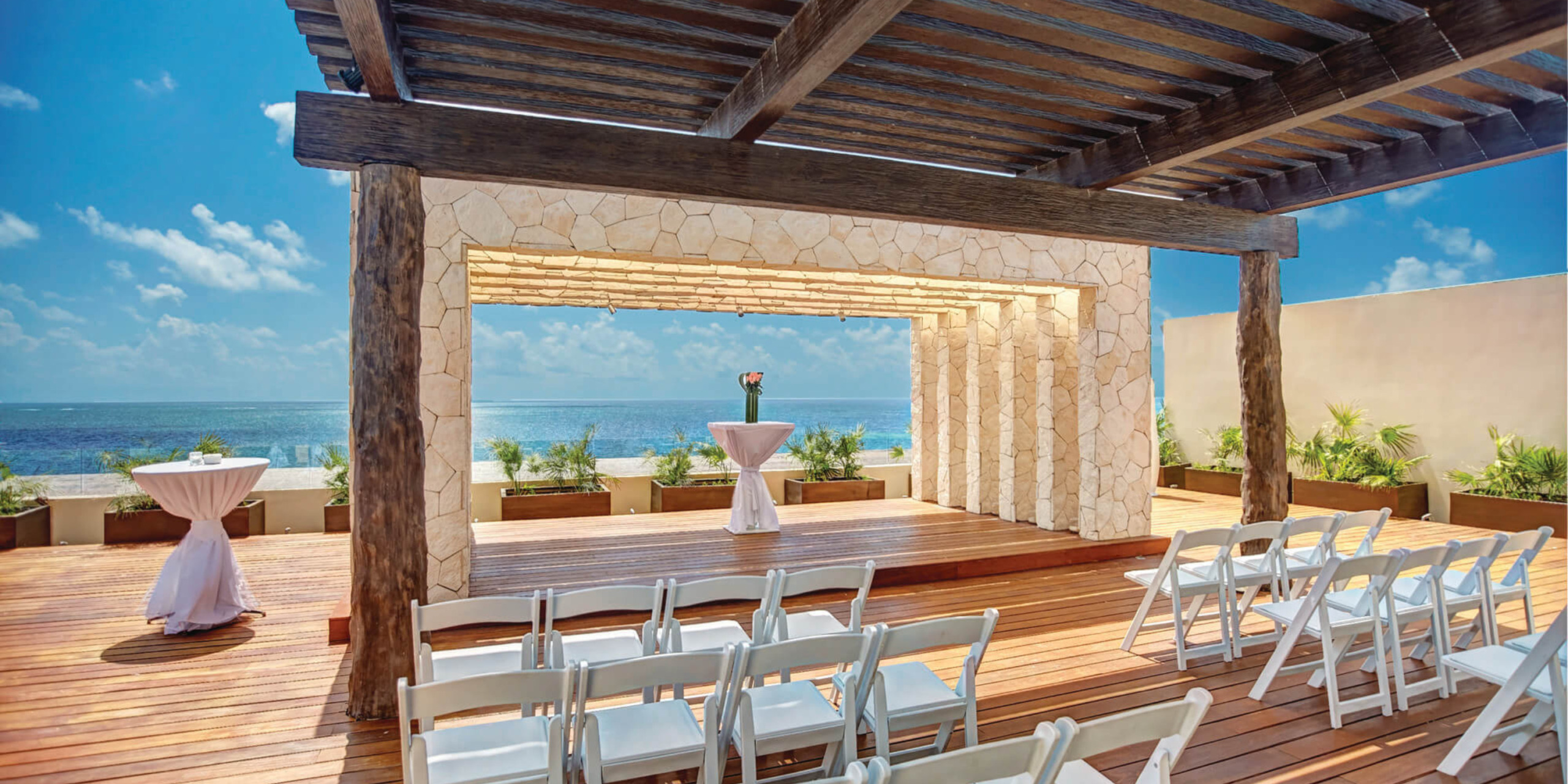 Destination wedding venue with a wooden pavilion and stone columns, overlooking the ocean, set up with white chairs and tables under a clear blue sky.