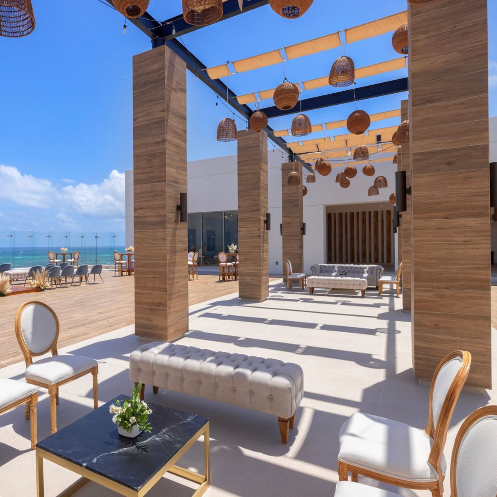A modern outdoor lounge designed for destination weddings features wooden accents, plush sofas, and enchanting hanging wicker lamps set against a backdrop of the clear blue sky.