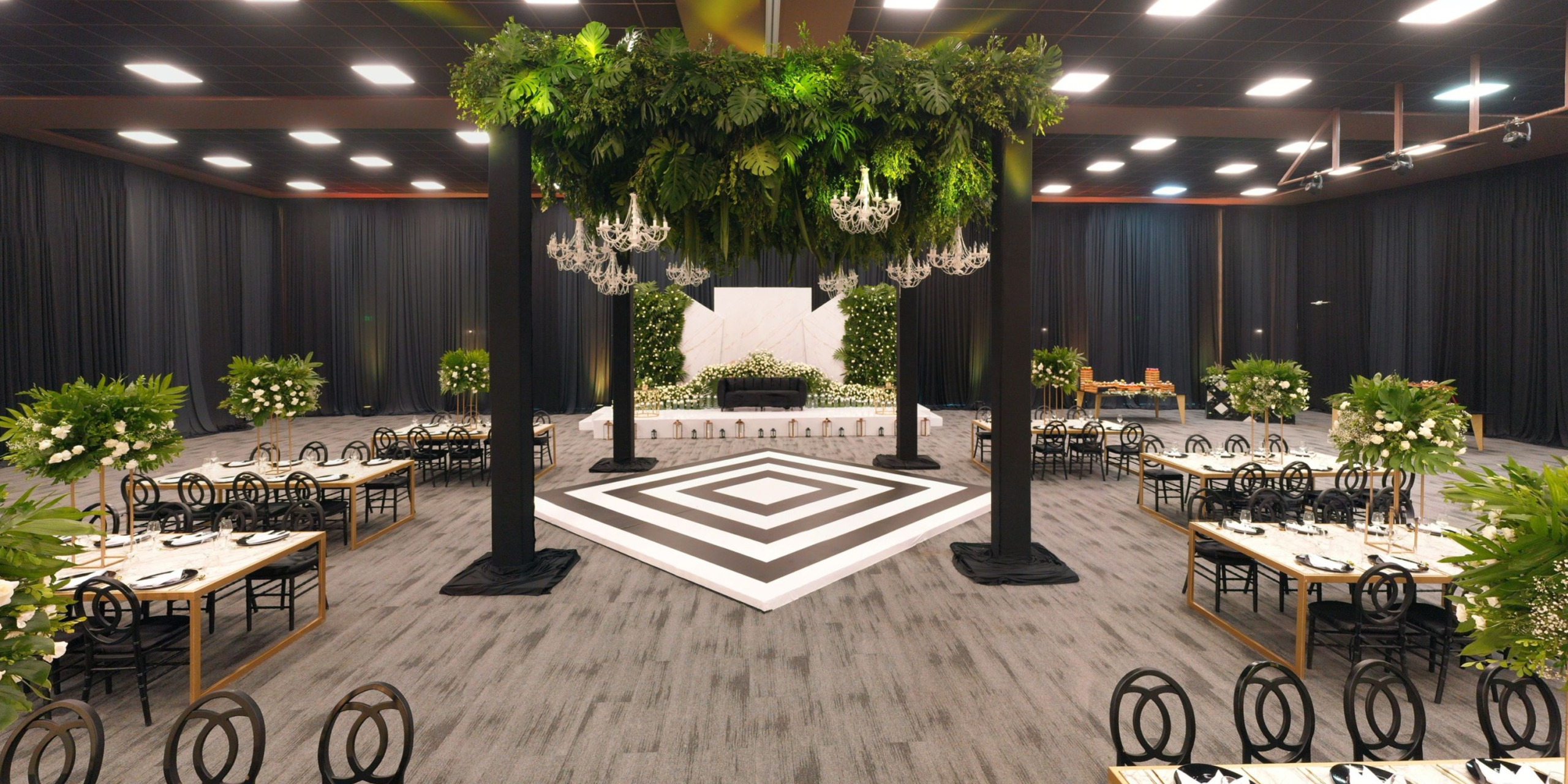 The Royalton Splash Riviera Cancun offers an elegant banquet hall featuring a black and white geometric dance floor, surrounded by dining tables with green floral arrangements, and complemented by suspended greenery decorating the ceiling.