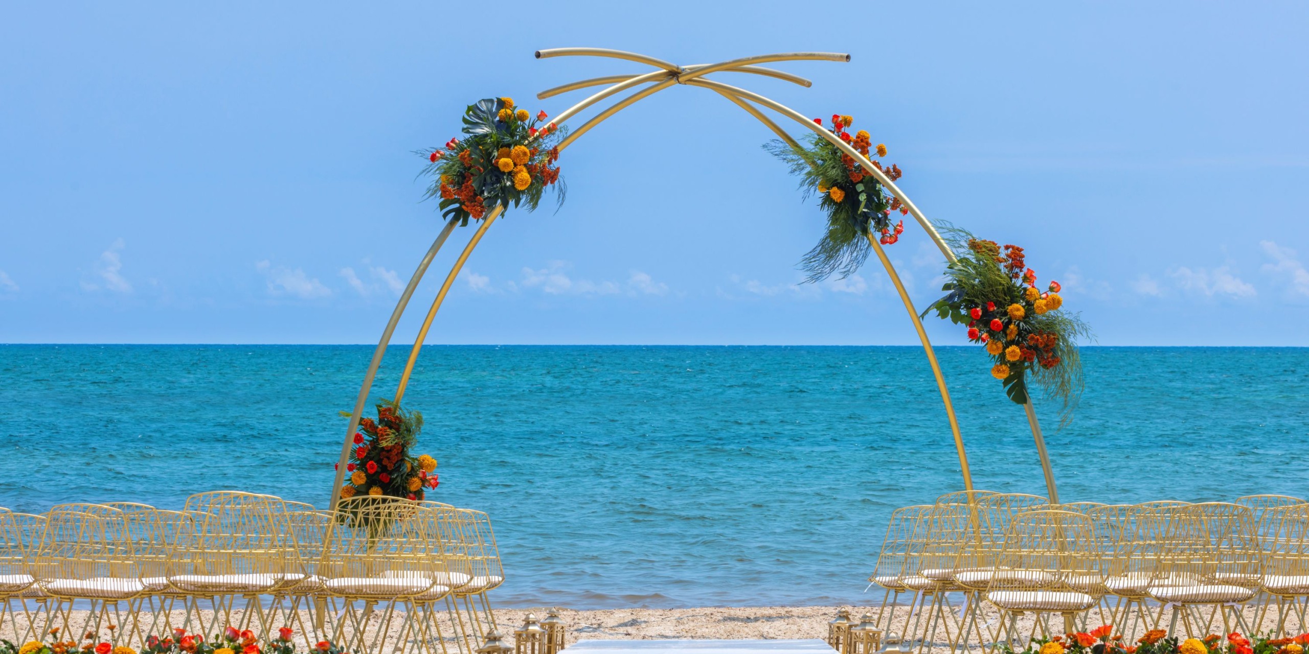 A wedding setup at the Royalton Splash Riviera Cancun featuring gold chairs and a floral-decorated arch, set against the backdrop of an ocean view beneath a clear blue sky.
