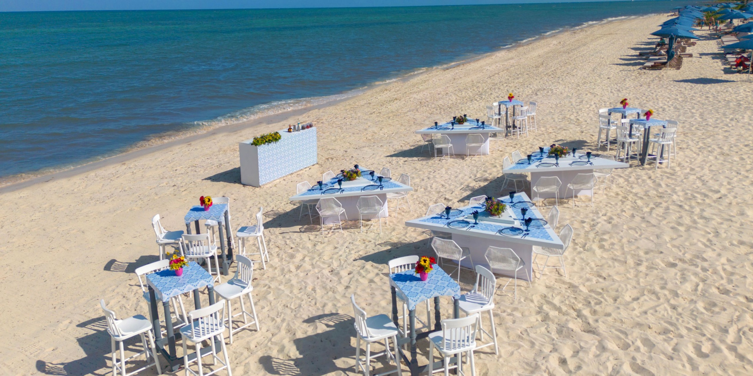 The Royalton Splash Riviera Cancun features dining tables adorned with blue covers and complemented by white chairs, perfectly situated on a sandy beach by the ocean, with a small bar in the background.