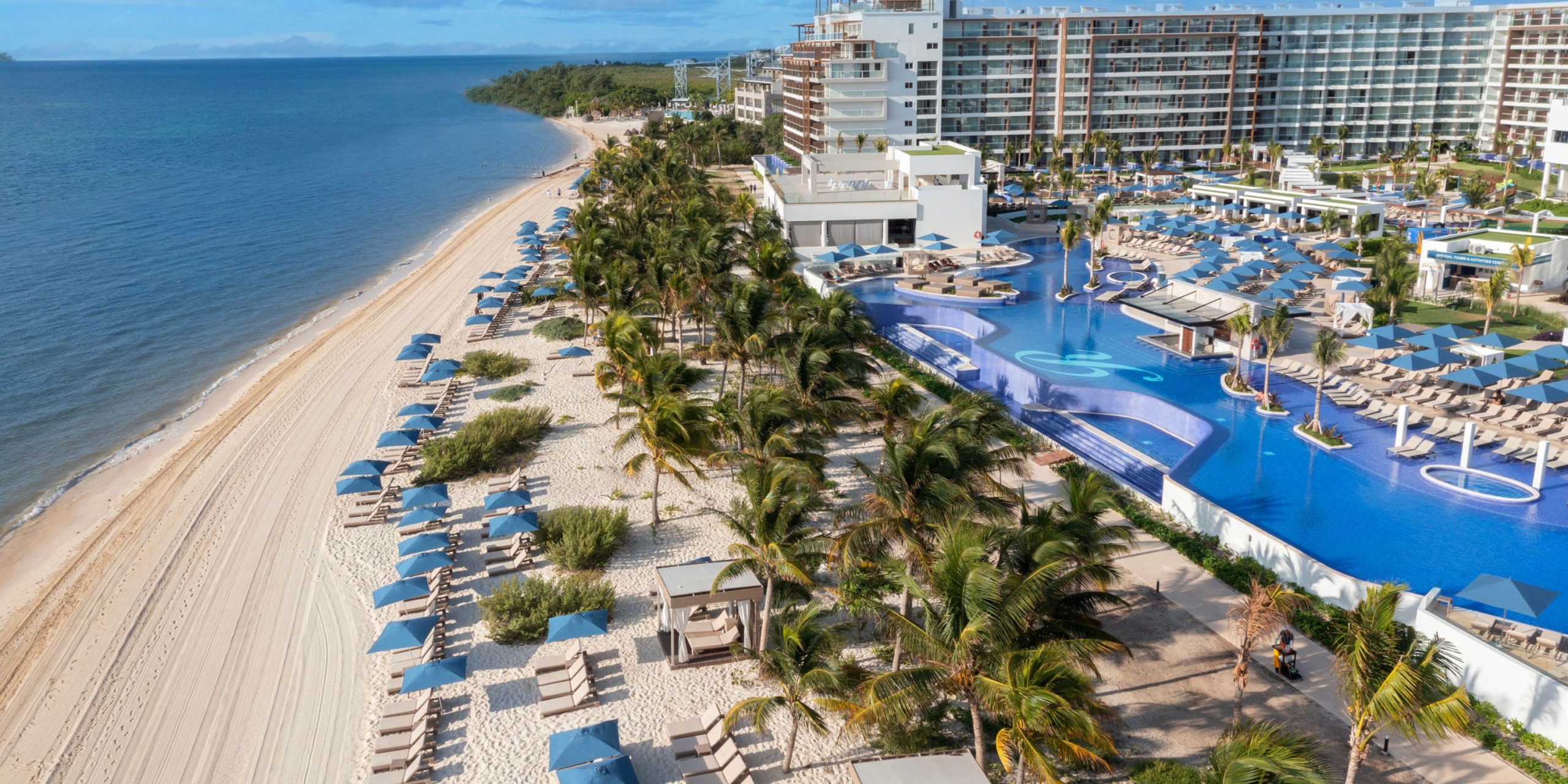 Aerial view of the Royalton Splash Riviera Cancun, a beachfront resort featuring a large outdoor pool framed by palm trees and lounge chairs under umbrellas, situated next to a sandy beach and the ocean.