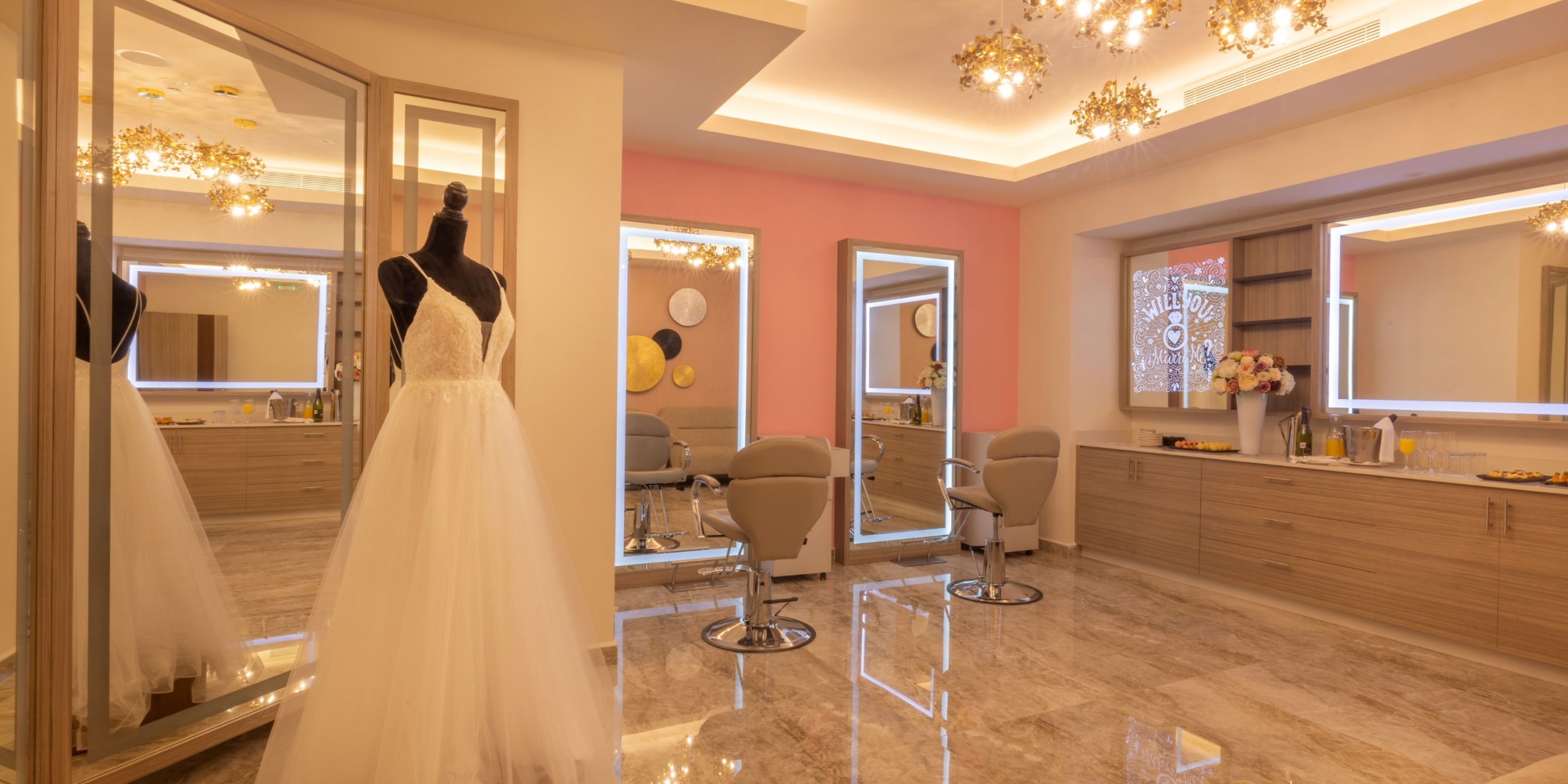 A showroom featuring a Royalton Splash Riviera Cancun white gown displayed on a mannequin, enhanced by a pink accent wall and complemented with mirrors and salon chairs. The room is adorned with marble flooring and decorative lighting.