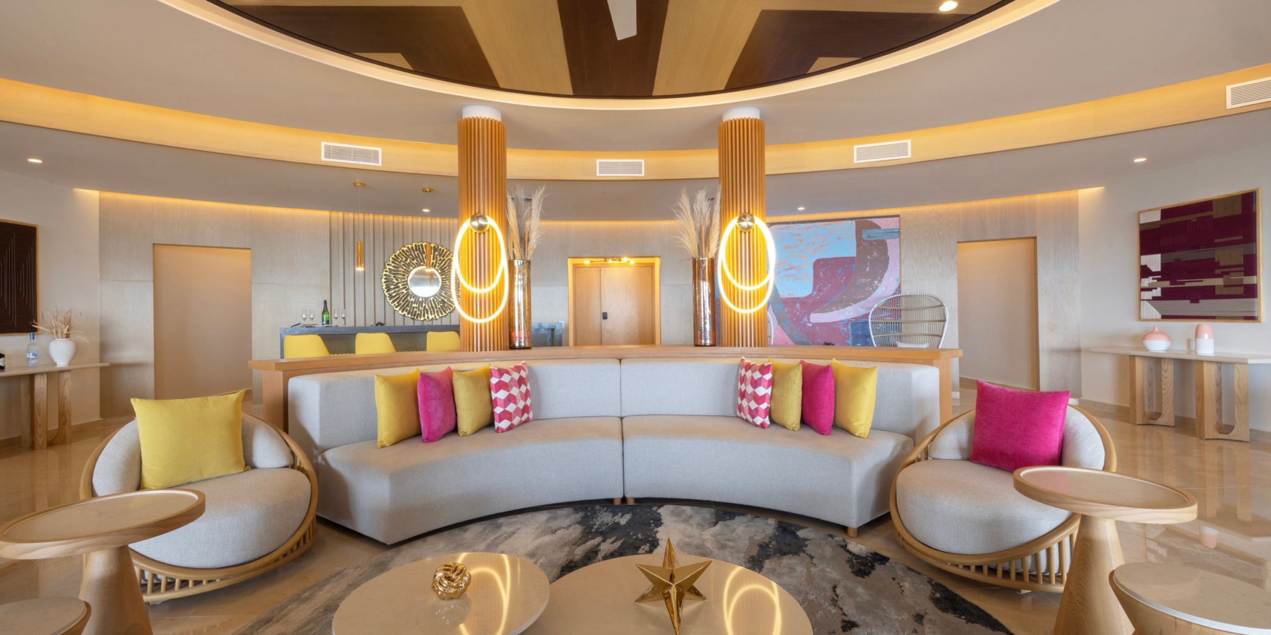 A contemporary lobby featuring the Royalton Splash Riviera Cancun's curved sofa, vibrant pillows, round tables adorned with decor, and artistic lighting in a circular room.