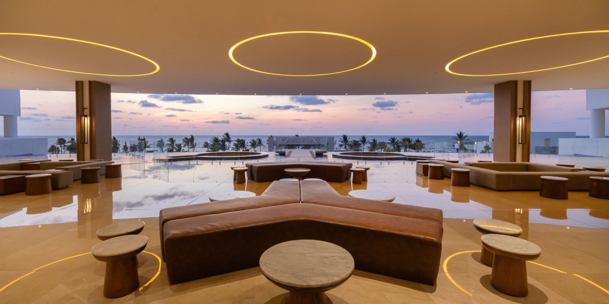 Experience a modern interior at Royalton Splash Riviera Cancun, featuring circular lights, a seating area, and tables that offer stunning views of the pool and ocean at sunset.