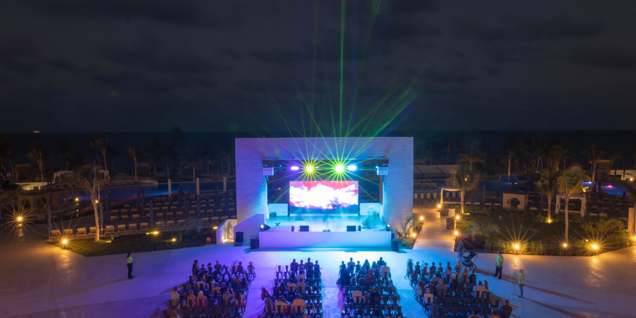 An evening outdoor concert at Royalton Splash Riviera Cancun, with a stage showcasing vibrant lights and a massive screen. The audience enjoys the performance while seated facing the stage, enveloped by palm trees and ambient warm lighting.