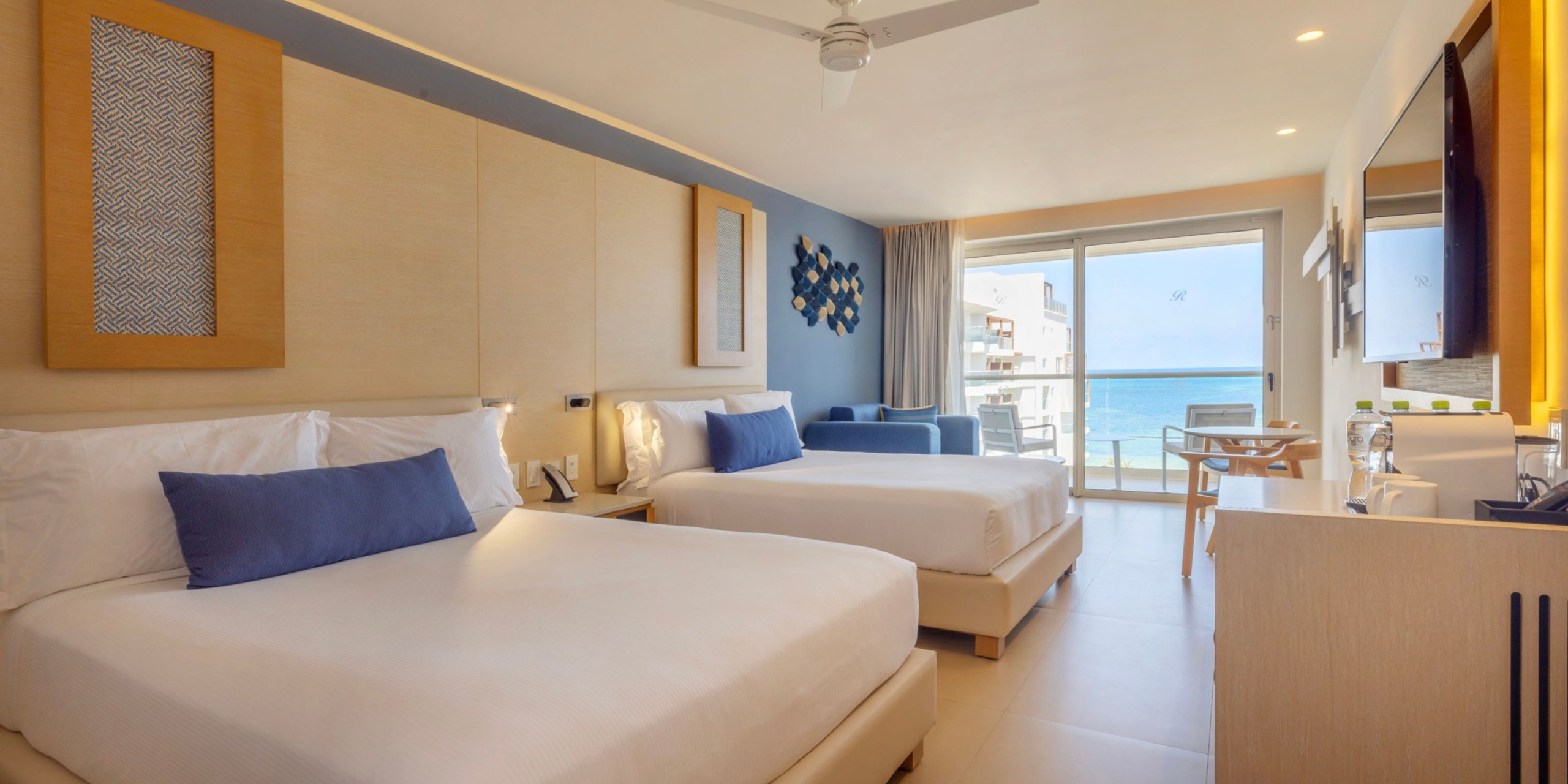Enjoy a stay at the Royalton Splash Riviera Cancun in a room featuring two double beds, elegant blue and white decor, a wall-mounted TV, a desk, a sofa, and breathtaking ocean views through a sliding glass door.