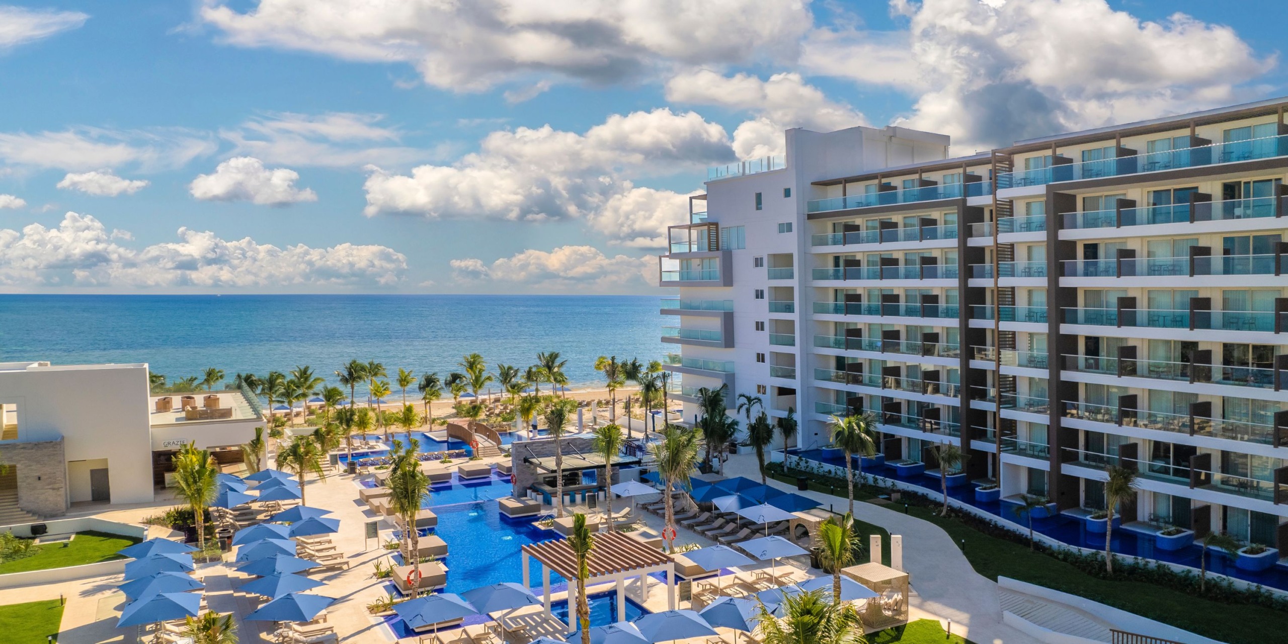The Royalton Splash Riviera Cancun features a large pool surrounded by palm trees and lounge chairs, situated next to a multi-story building by the ocean under a partly cloudy sky.