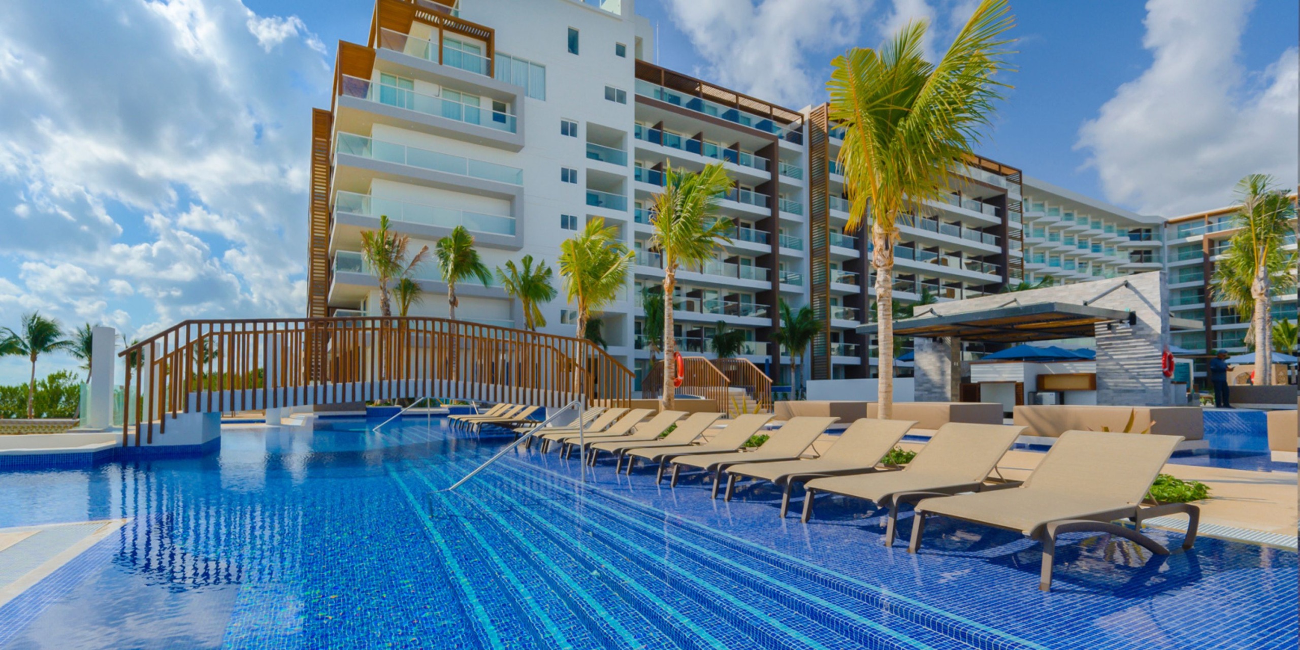 Relax by the outdoor pool at Royalton Splash Riviera Cancun, surrounded by lounge chairs and palm trees, with the modern multi-story hotel providing a stunning backdrop under a partly cloudy sky.