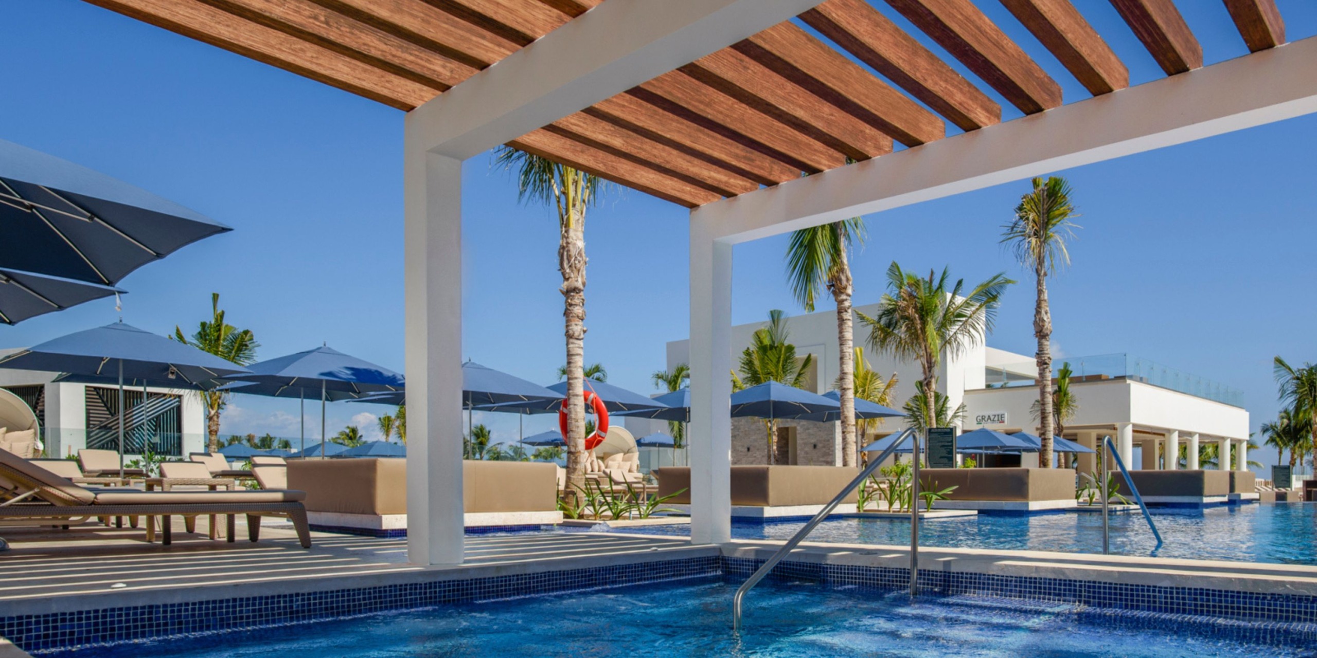 The poolside area at the Royalton Splash Riviera Cancun features blue umbrellas, sun loungers, and palm trees. A wooden pergola offers shade over a section of the pool, with a building visible in the background.