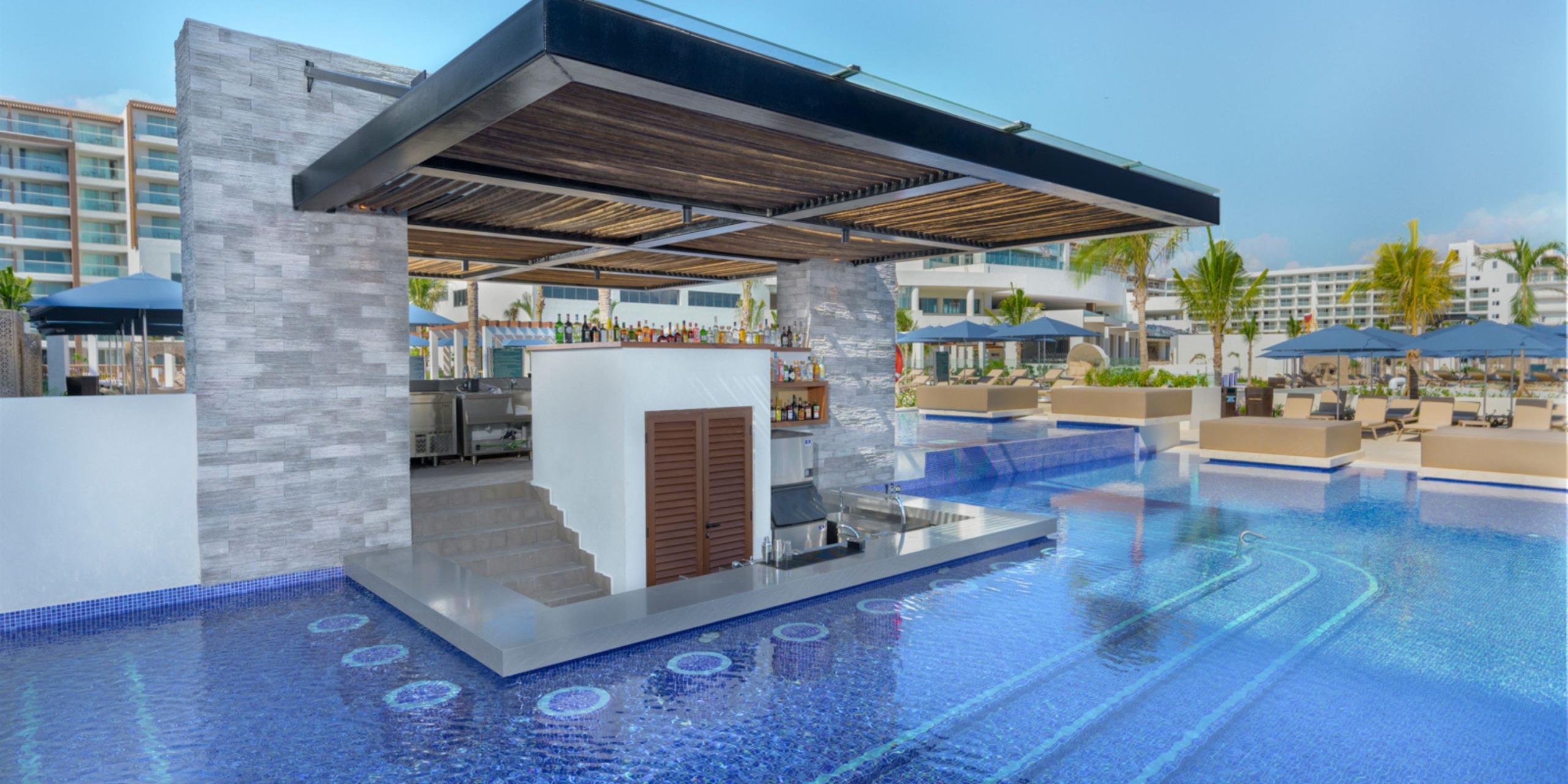 The Royalton Splash Riviera Cancun features an outdoor swim-up bar with stools in a pool, surrounded by resort buildings and lounge chairs shaded by umbrellas.