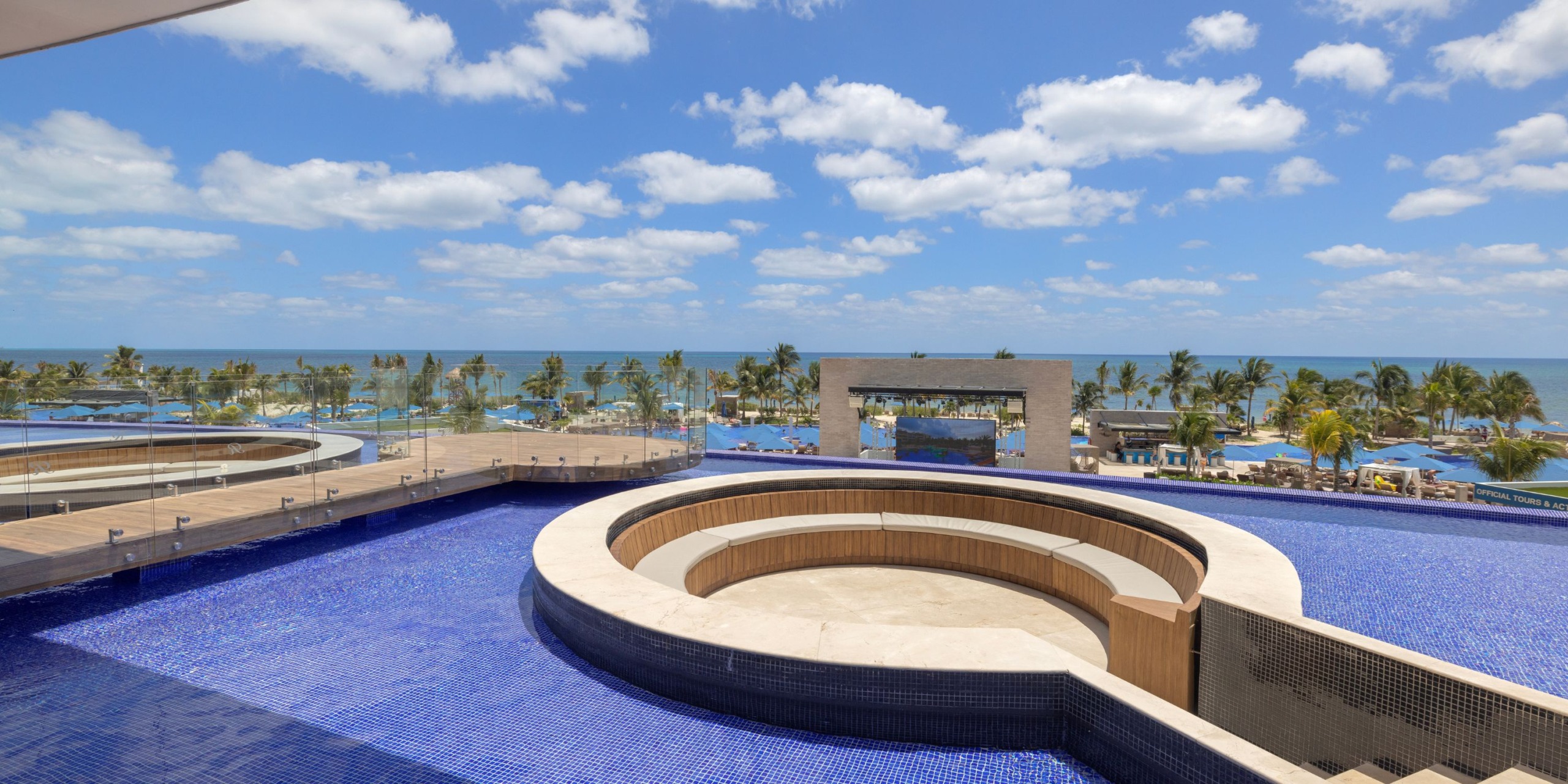 The Royalton Splash Riviera Cancun features a rooftop infinity pool with a circular seating area, offering stunning views of palm trees and the ocean beneath a blue sky dotted with scattered clouds.