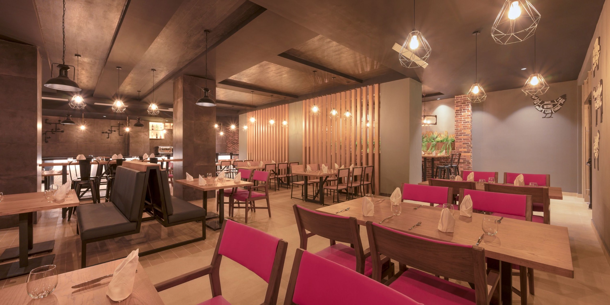 The interior of Royalton Splash Riviera Cancun showcases a contemporary design with wooden tables, pink cushioned chairs, and pendant lights. The space is beautifully accented with wooden partitions and brick walls.