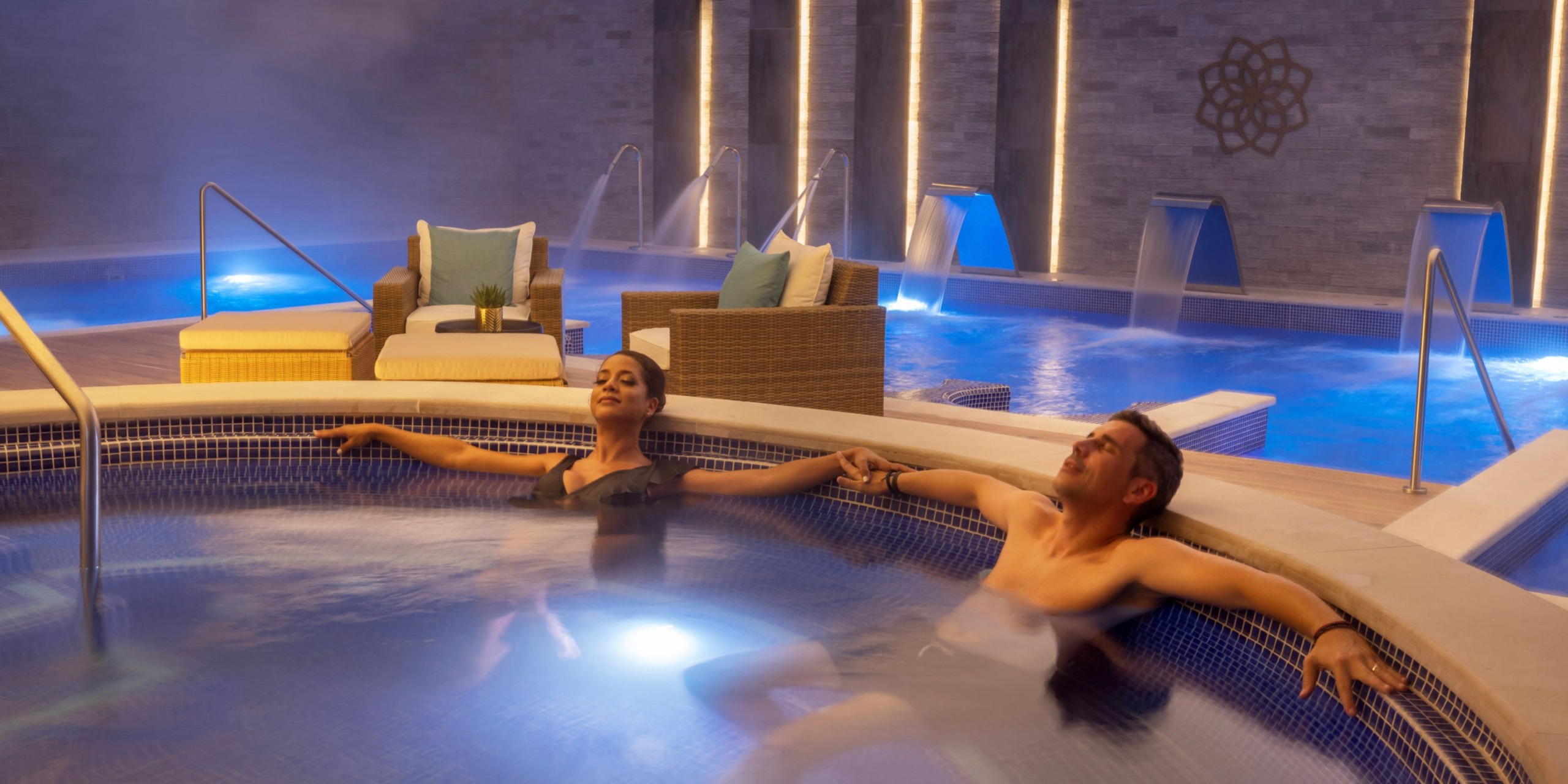 Two people relax in a dimly-lit indoor pool area at Royalton Splash Riviera Cancun, featuring lounge chairs and illuminated water features in the background.