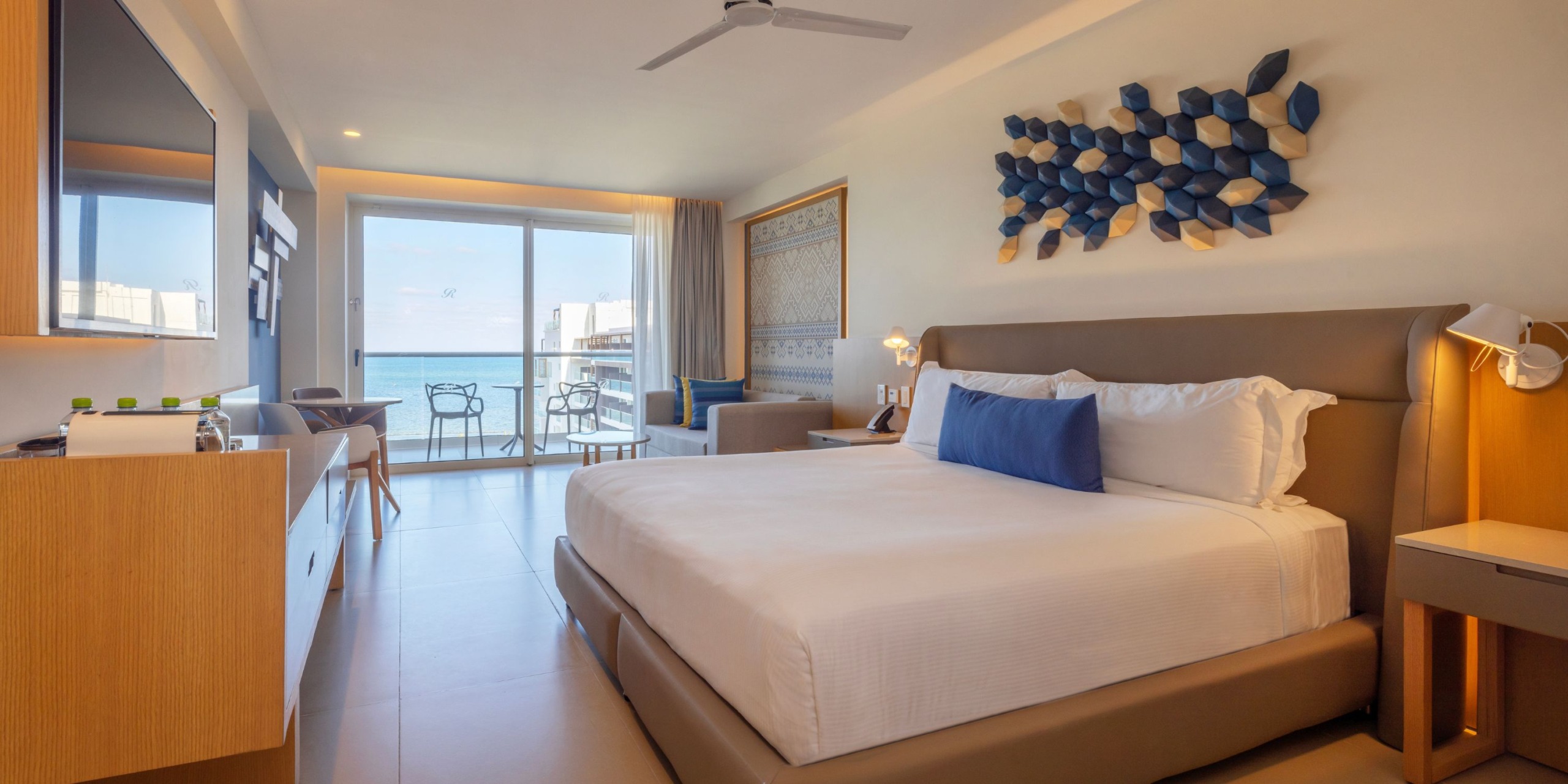Experience a stay at the Royalton Splash Riviera Cancun in a hotel room featuring a large bed and blue and white decor. Enjoy amenities like a flat-screen TV and a balcony with breathtaking ocean views. The modern furnishings include a desk, chair, and wall art adorned with 3D hexagonal shapes.