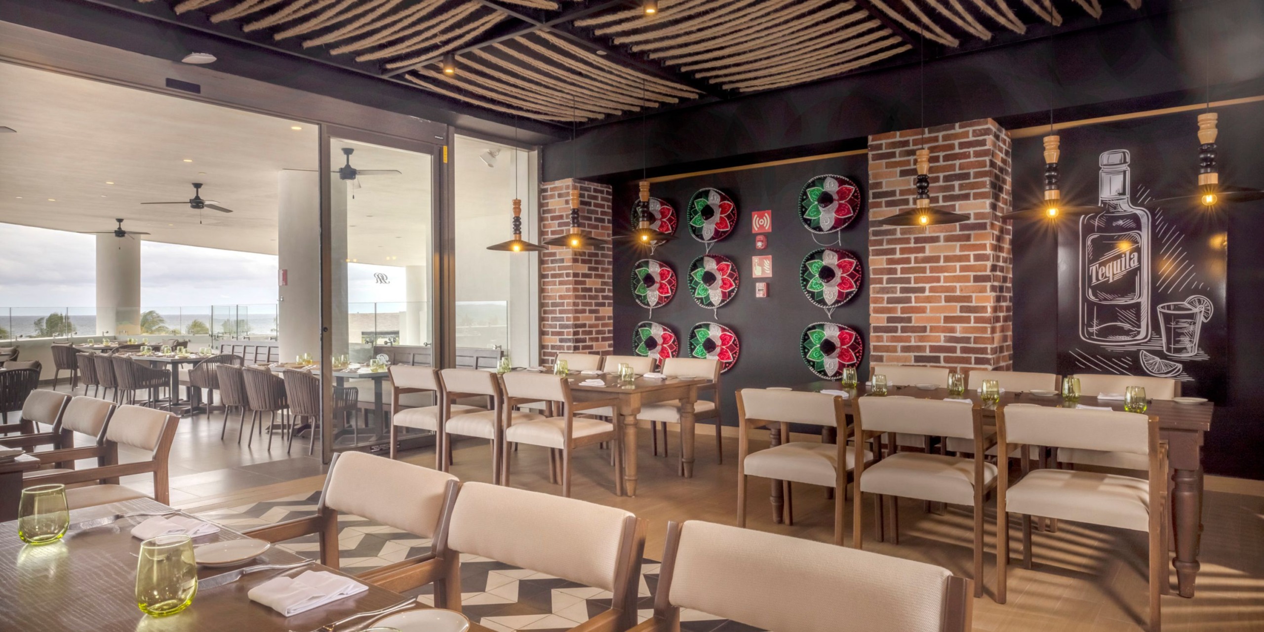 The interior of the Royalton Splash Riviera Cancun features a contemporary design with wooden furniture, exposed brick walls, intricate chalkboard art, and stylish decorative lighting. The expansive windows open up to reveal an outdoor dining area offering scenic views.