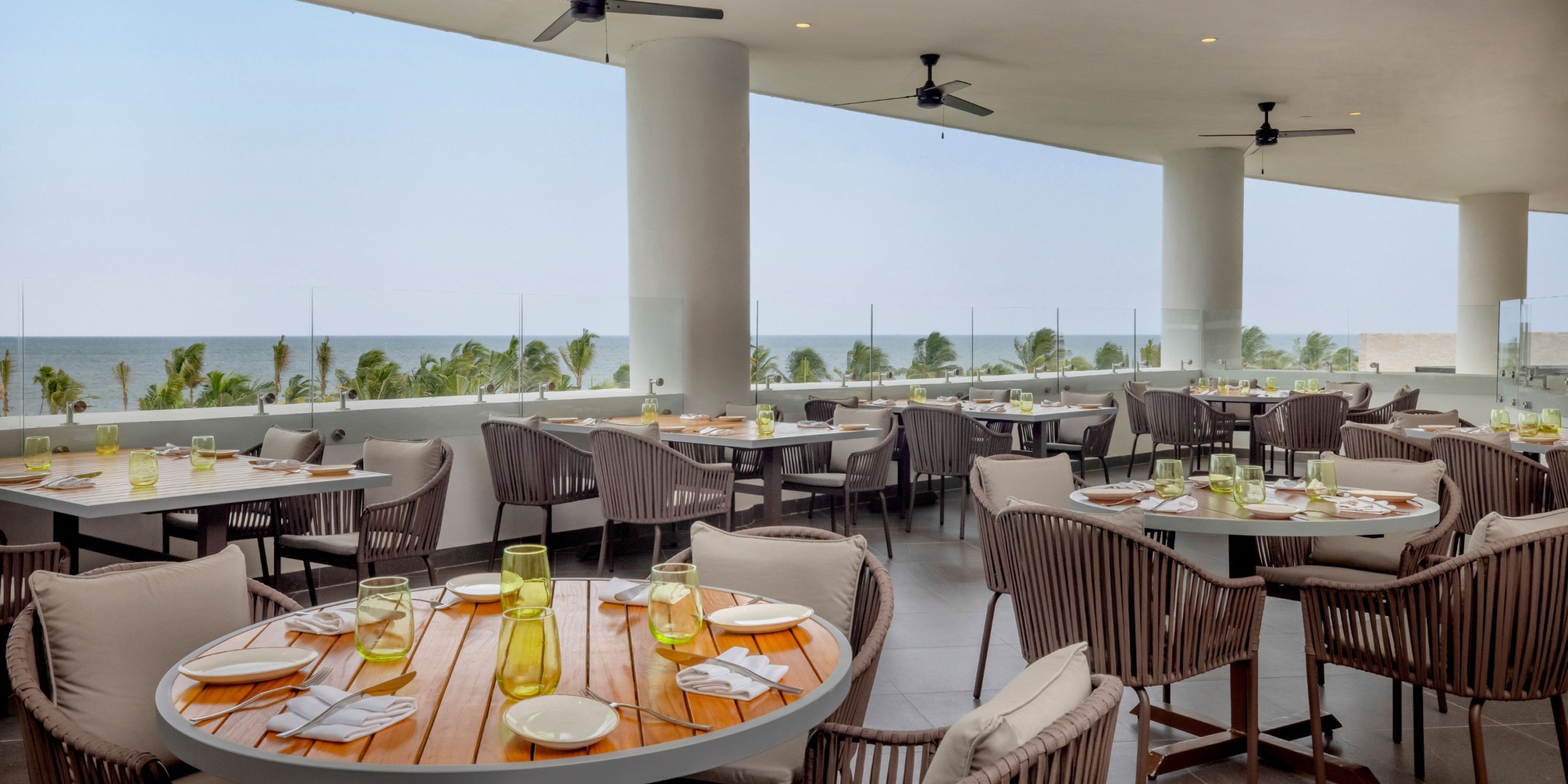 The outdoor dining area at the Royalton Splash Riviera Cancun features wooden tables adorned with clear glasses and white napkins, offering a scenic view of palm trees and the ocean in the background, with ceiling fans overhead.