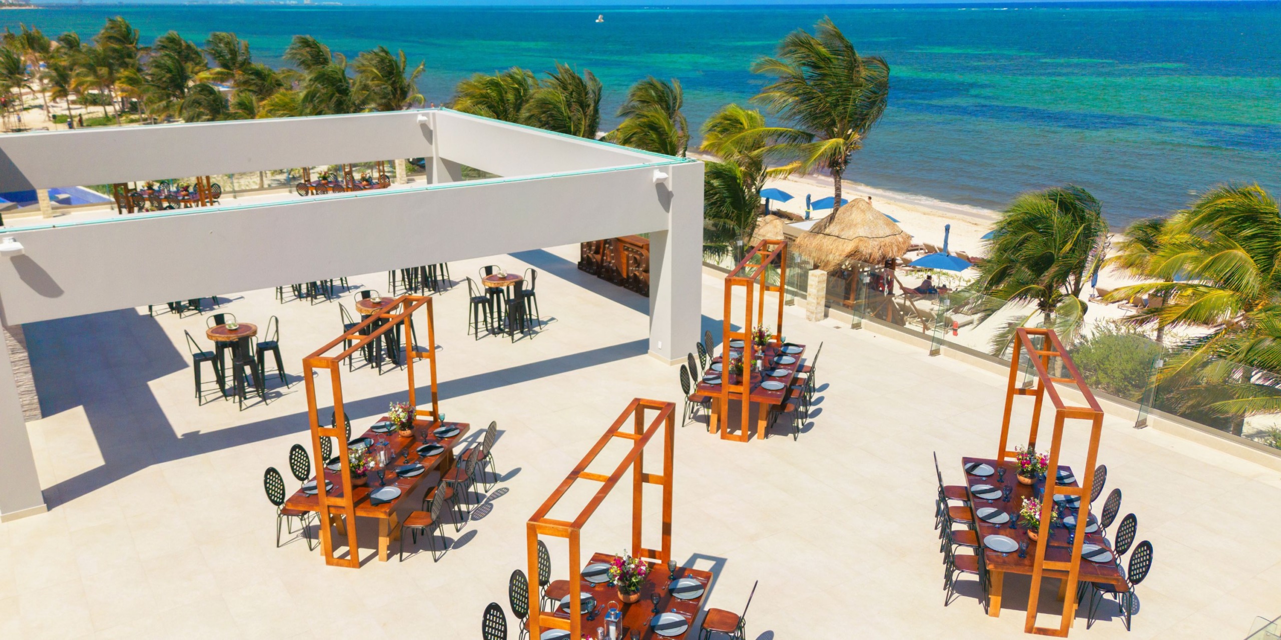 The outdoor dining area at Royalton Splash Riviera Cancun features neatly arranged tables and chairs, surrounded by palm trees and an expansive view of the clear blue ocean.