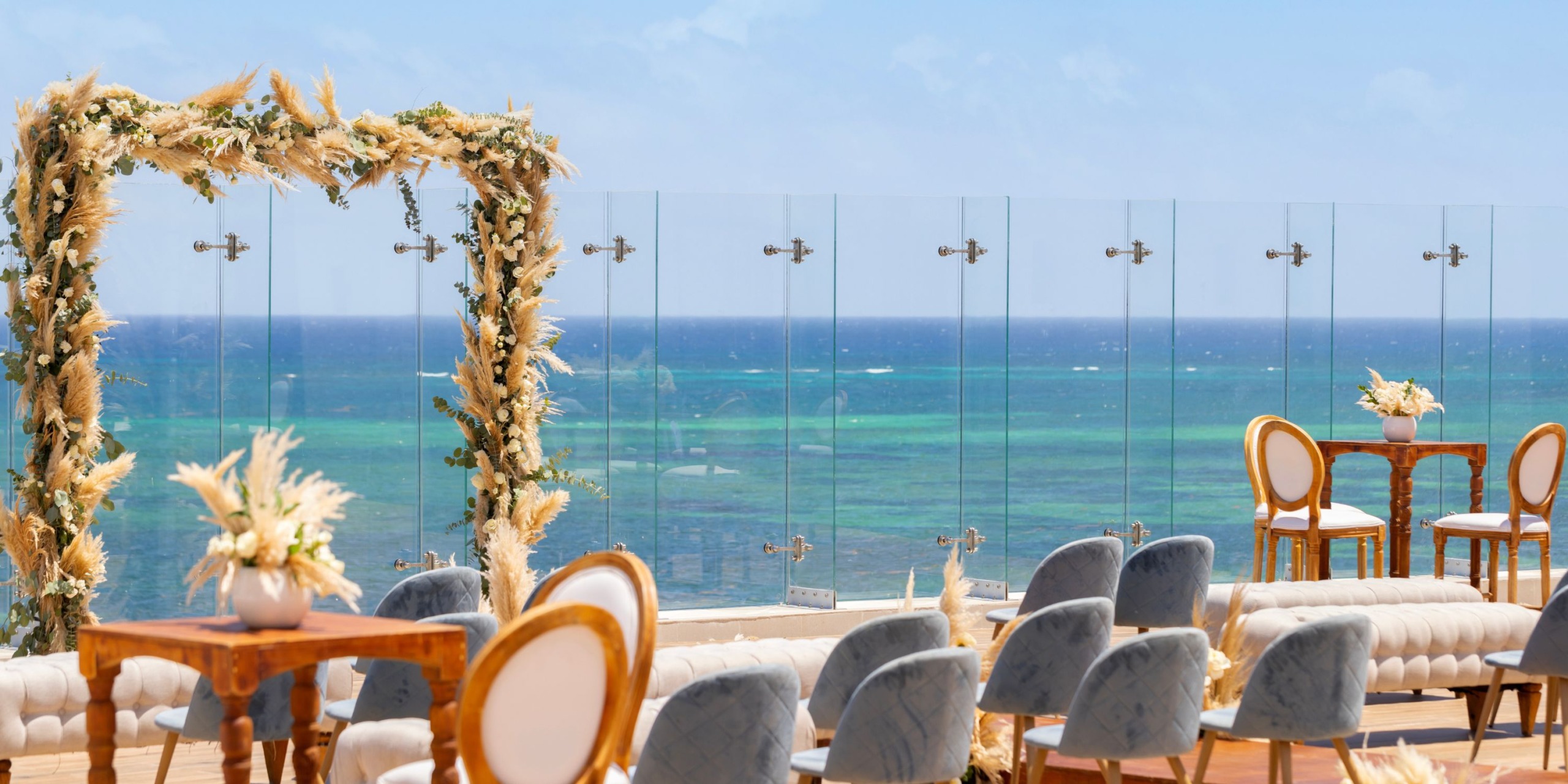 Experience the Royalton Splash Riviera Cancun's outdoor wedding setup with elegant chairs and tables on a terrace overlooking the ocean, accented by a floral arch and clear glass railing.