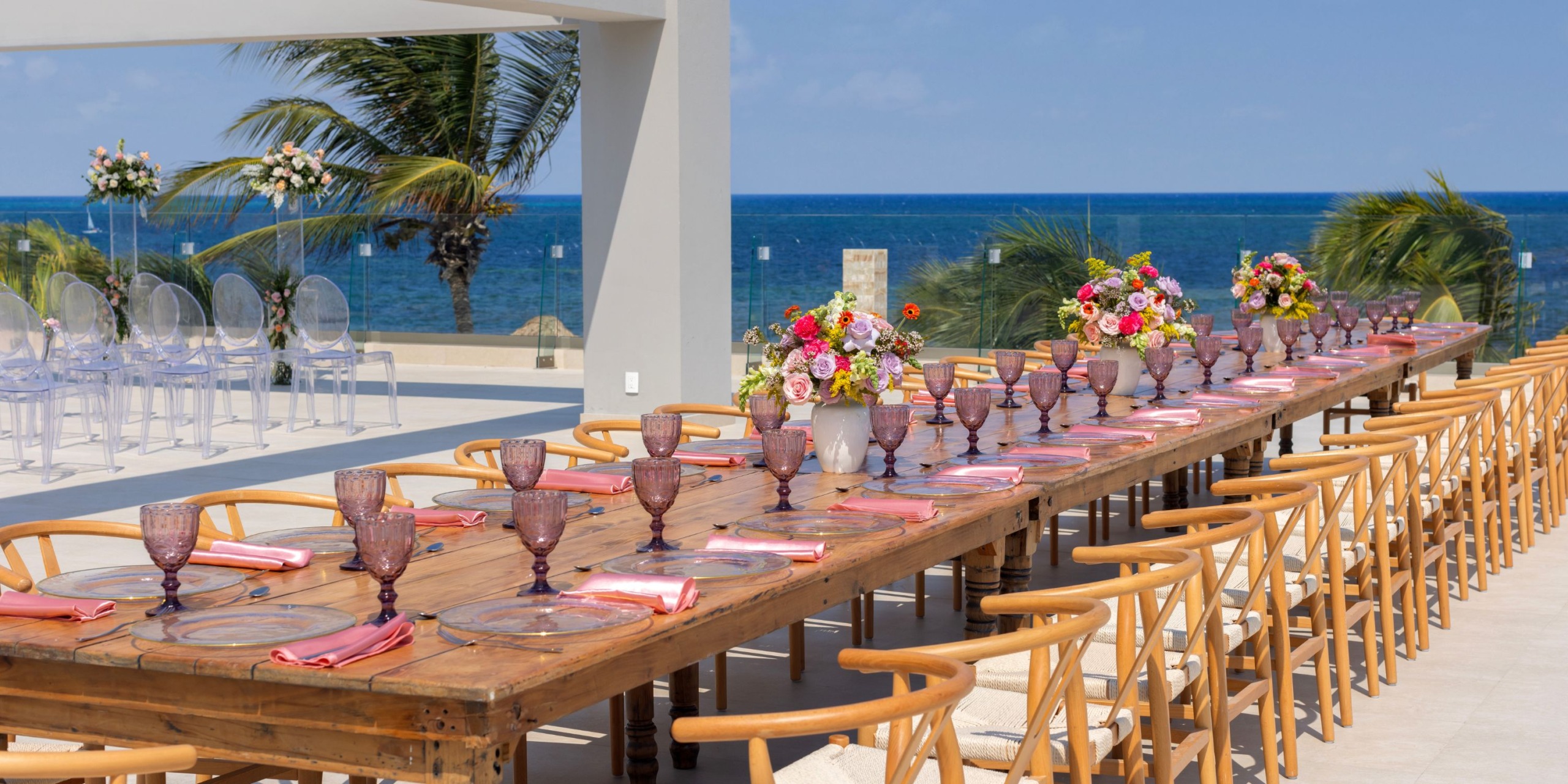 A luxurious setup at the Royalton Splash Riviera Cancun features a long wooden table adorned with pink napkins and elegant purple glassware. The table is beautifully enhanced by vibrant flower arrangements, all set against the backdrop of an ocean view and towering palm trees.