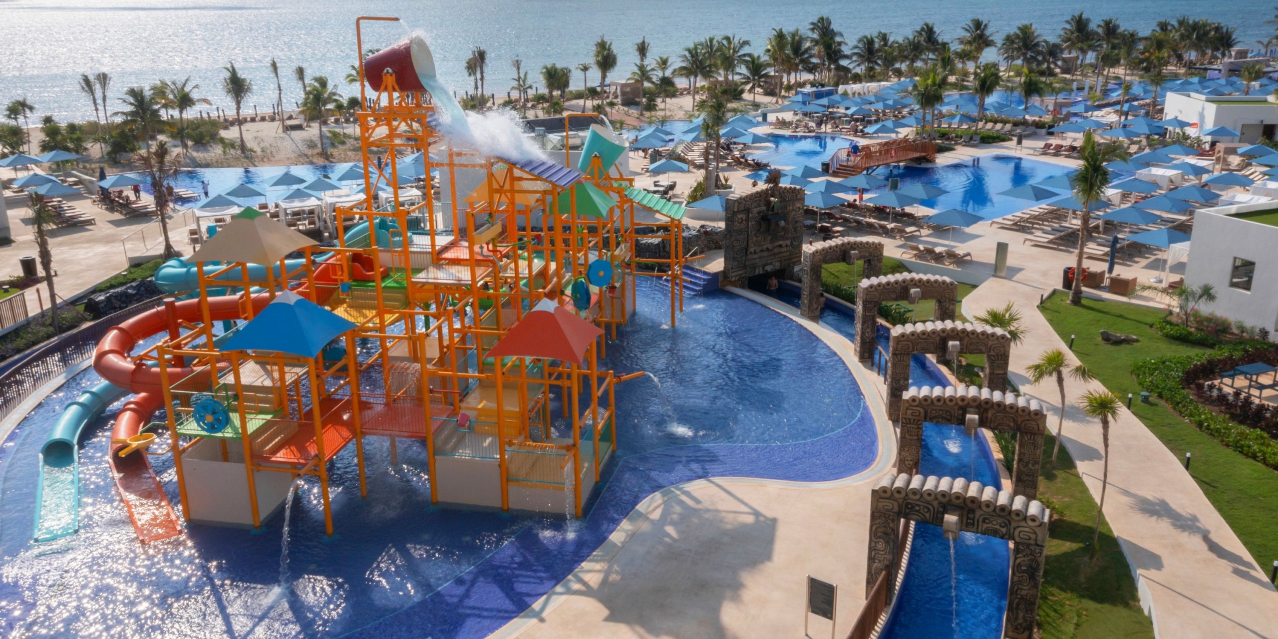 The Royalton Splash Riviera Cancun features a large water playground with vibrant structures and slides, all encompassed by a spacious pool area. In the background near the coast, palm trees and lounge chairs can be seen.