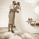 A bride and groom share a kiss on a sandy beach at sunset during their destination wedding, framed by gentle waves and a clear sky.