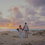 A couple in wedding attire holding hands on a beach at sunset with clouds in the sky during their destination wedding.