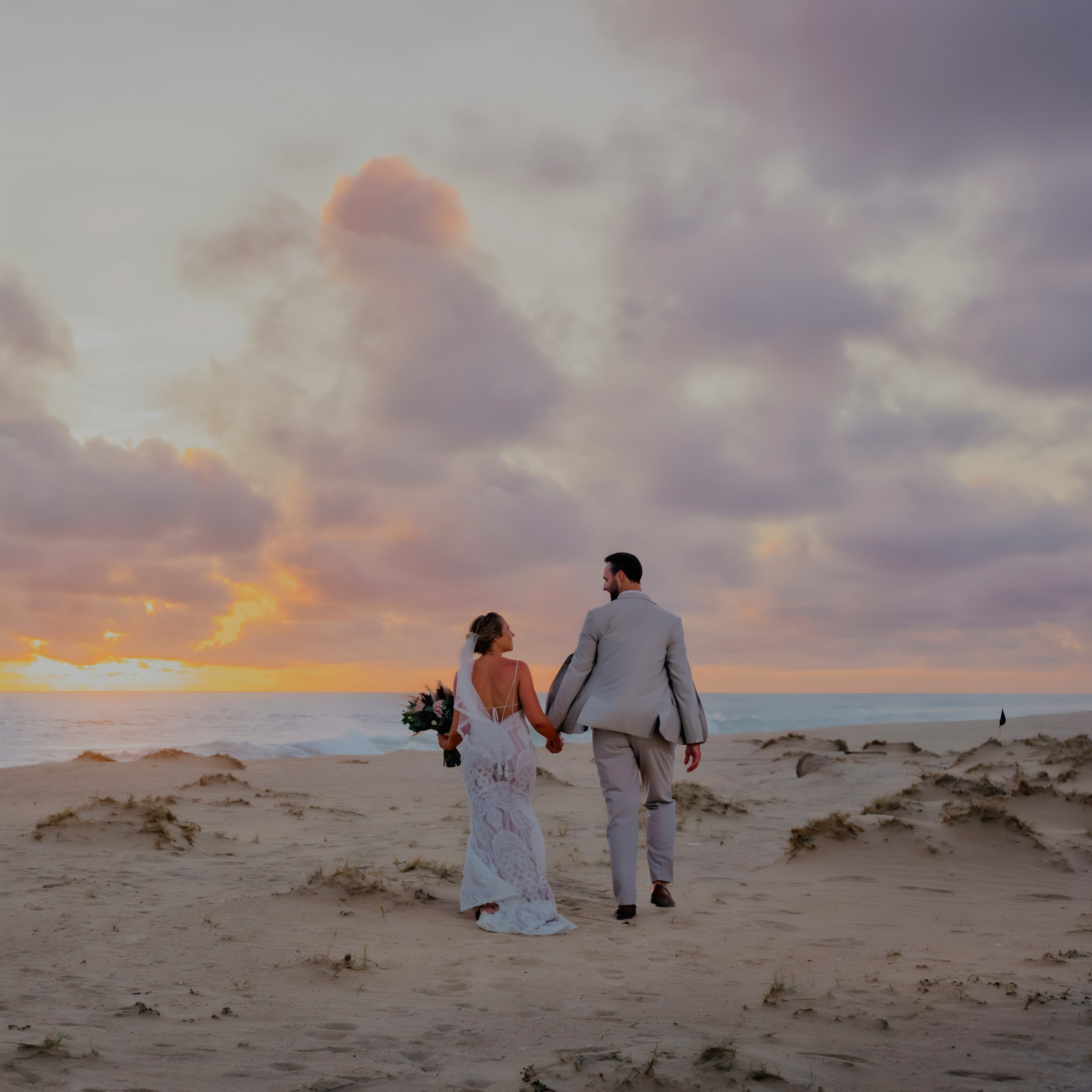 A couple in wedding attire holding hands on a beach at sunset with clouds in the sky during their destination wedding.
