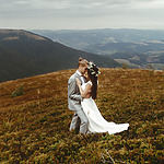 A couple in destify wedding attire embraces on a mountain, with sweeping views of the valley and overcast sky in the background.