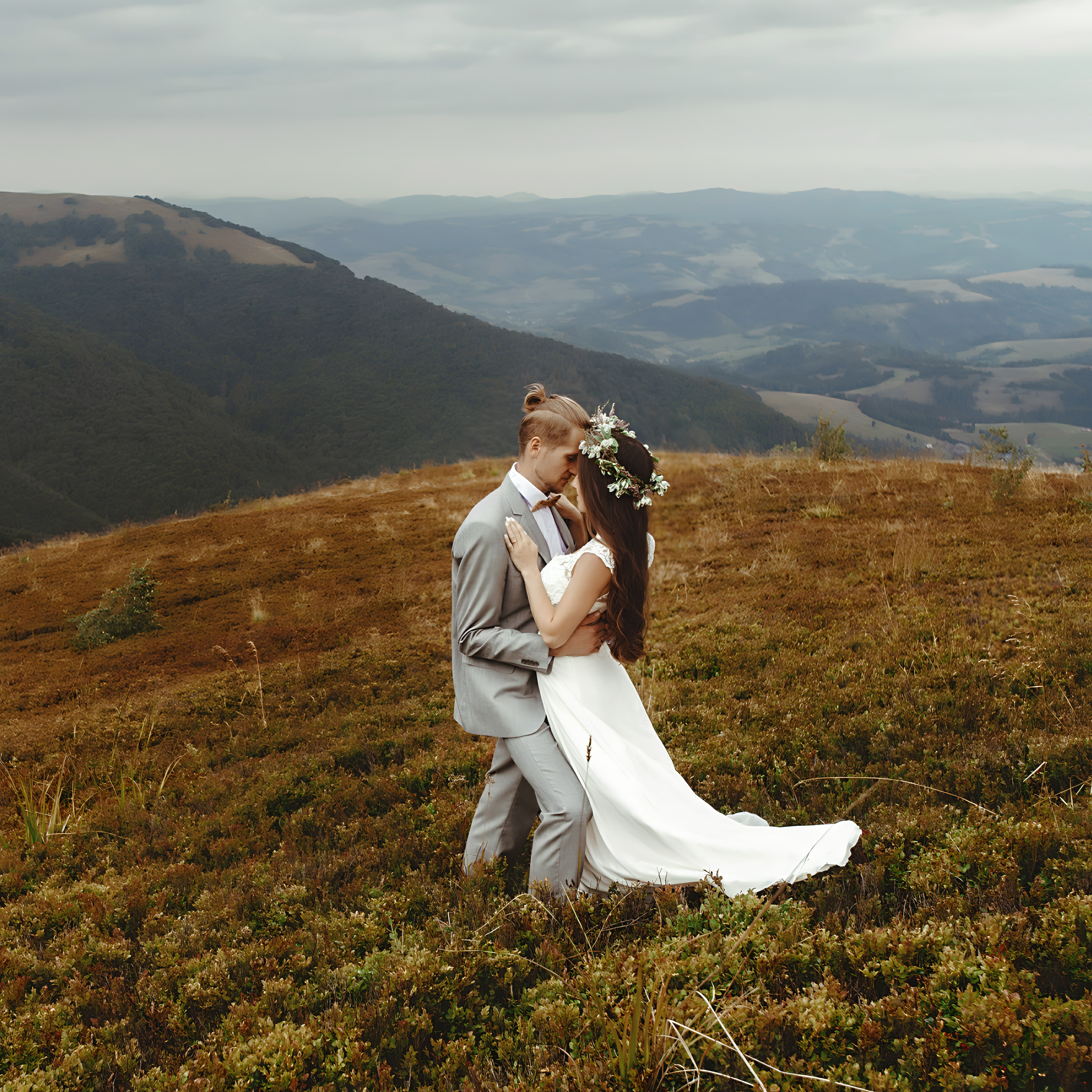 A couple in destify wedding attire embraces on a mountain, with sweeping views of the valley and overcast sky in the background.