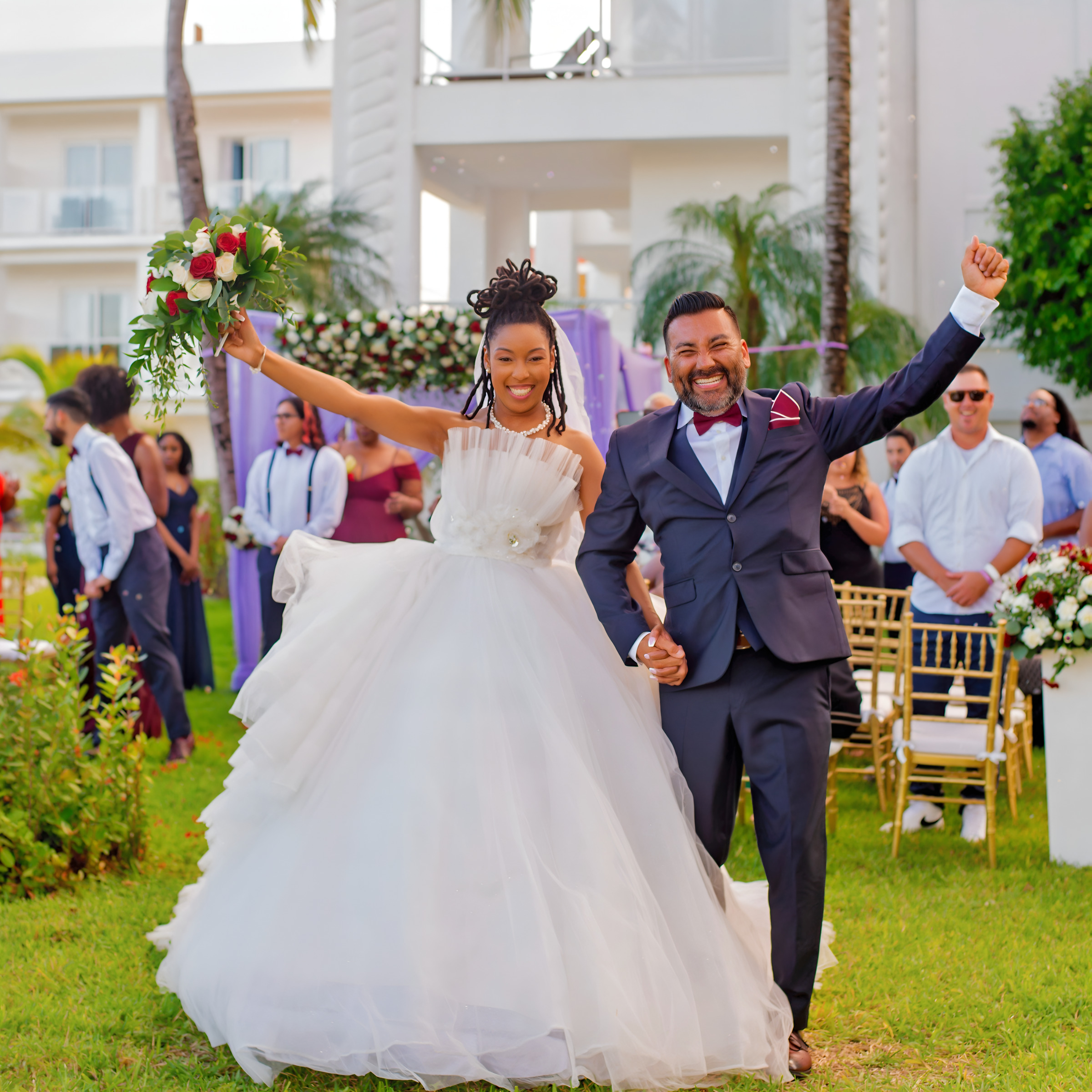 A joyful bride and groom walking hand in hand at their destination wedding, the bride holding a bouquet, guests in the background.