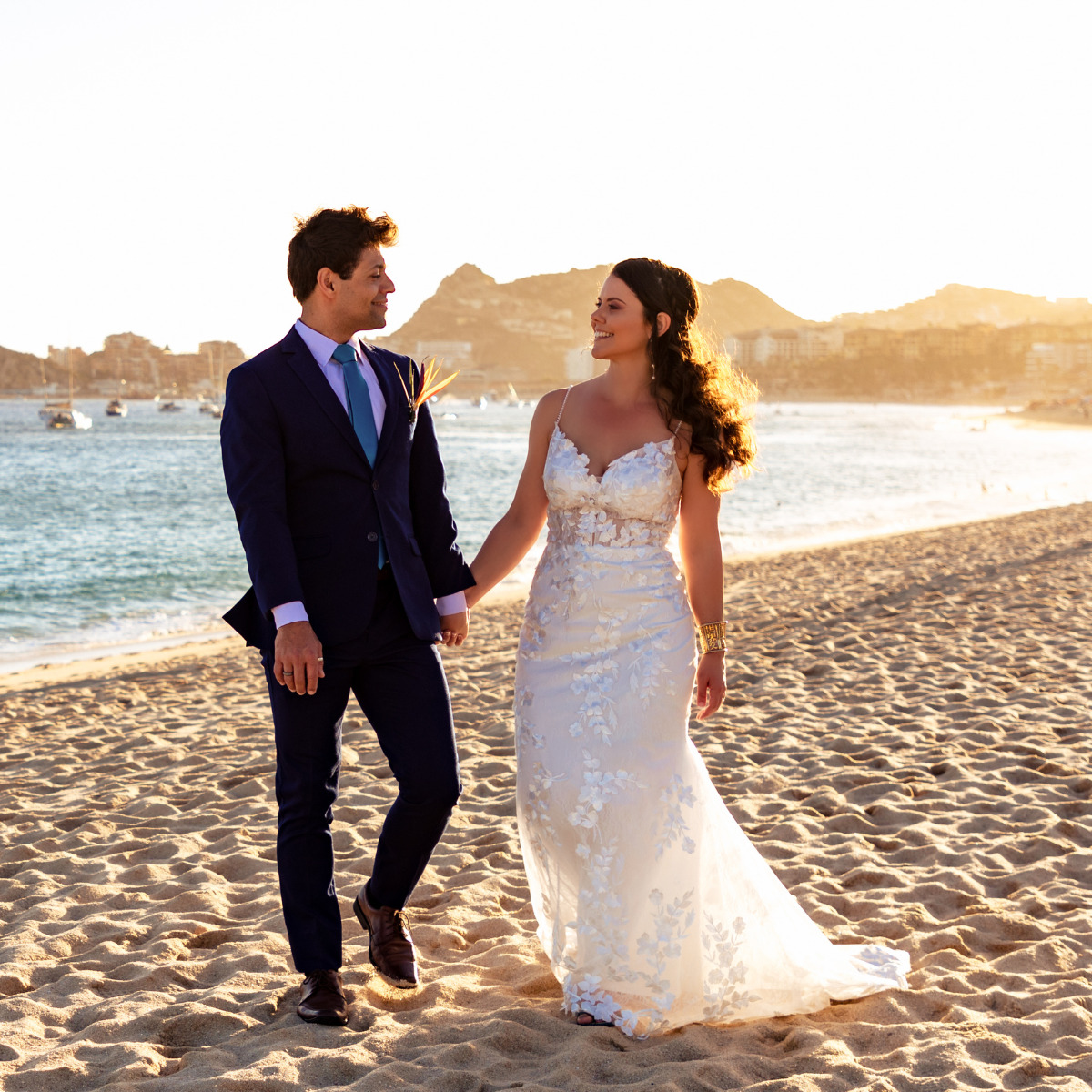 A newlywed couple in formal attire walks hand-in-hand on a sandy beach at sunset, with boats and rocks in the background, at one of the top destination wedding locations.