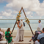 A couple kisses under a triangular floral arch during a perfect destination wedding ceremony on the beachfront, with guests seated and an officiant standing by.