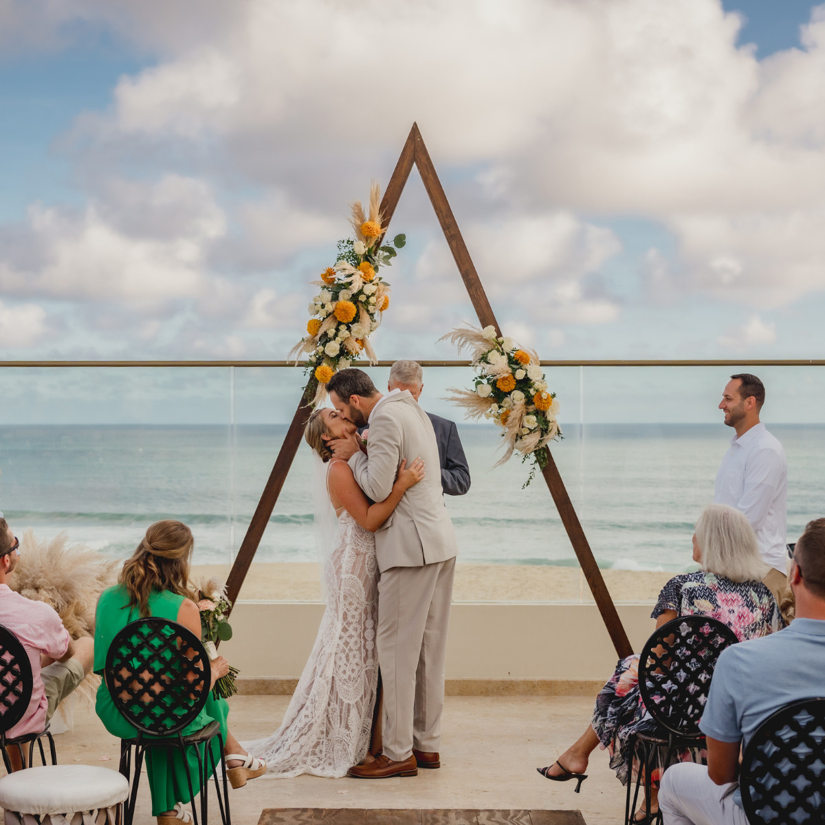 A couple kisses under a triangular floral arch during a perfect destination wedding ceremony on the beachfront, with guests seated and an officiant standing by.